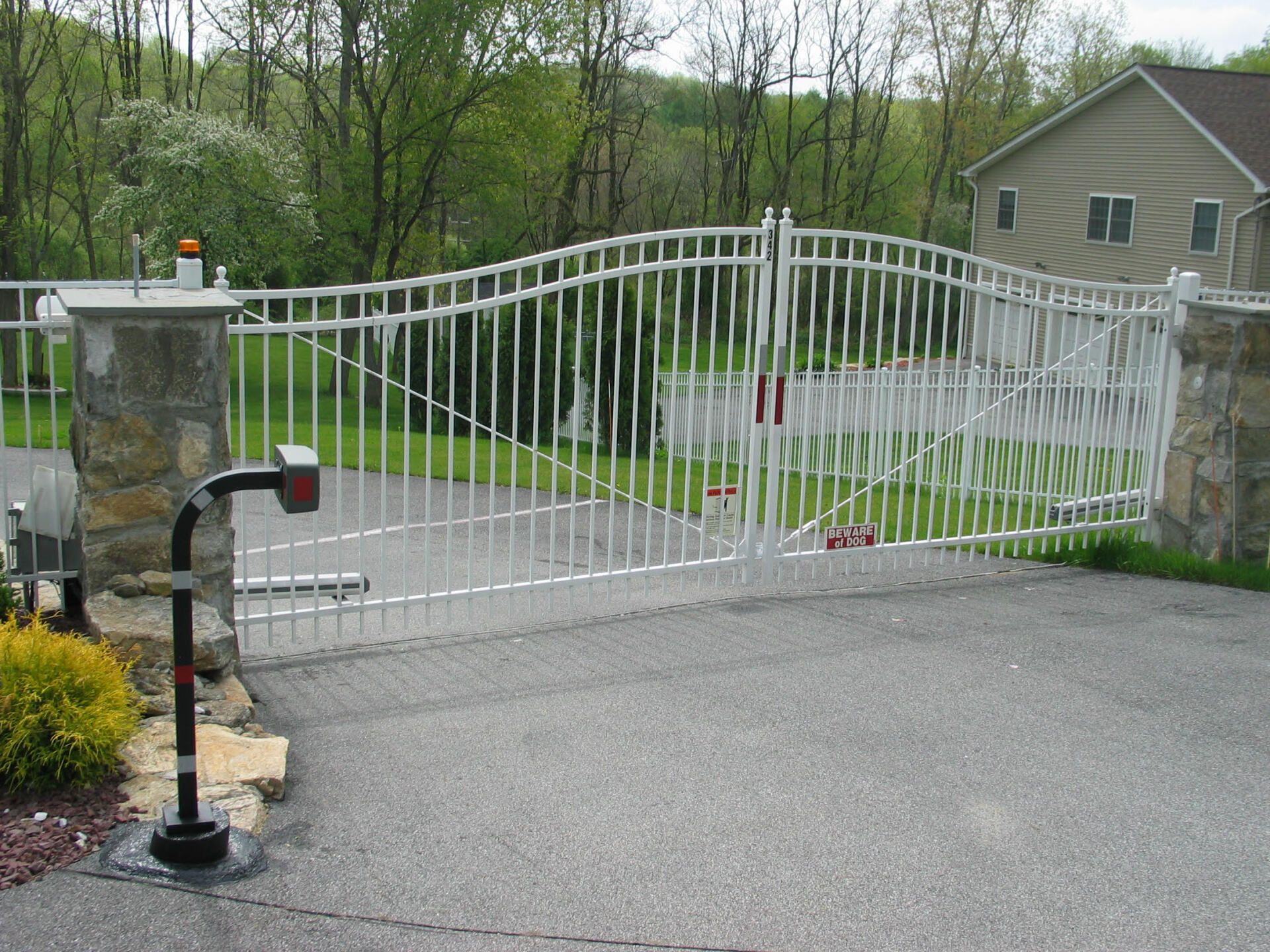 White metal driveway gate across a paved entrance, with stone pillars and greenery behind it