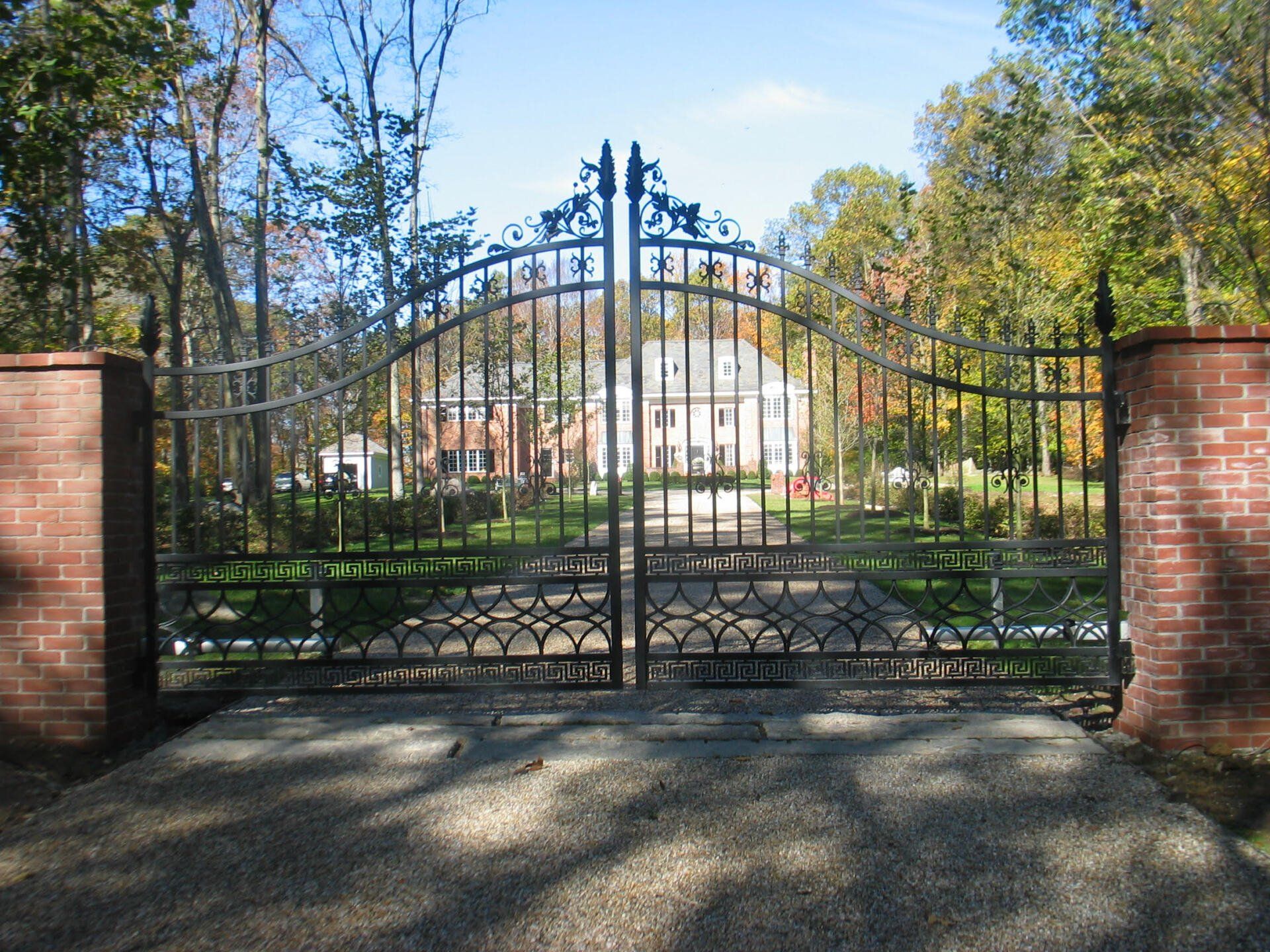 Wrought-iron gate between brick pillars in front of a large white building and trees under a blue sky
