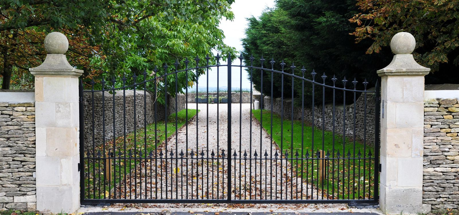 White metal driveway gate across a paved driveway beside a house and stone post in a suburban yard
