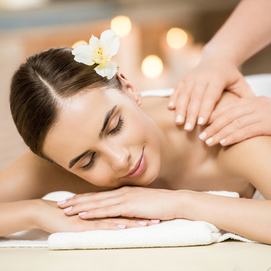 Una donna riceve un massaggio in una spa con un fiore bianco tra i capelli, sdraiata su asciugamani piegati in una stanza illuminata da luci soffuse.