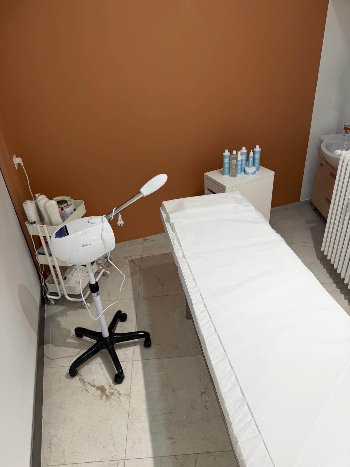Piccola stanza clinica con lettino per trattamenti, sgabello con rotelle e attrezzature mediche bianche su una parete arancione.