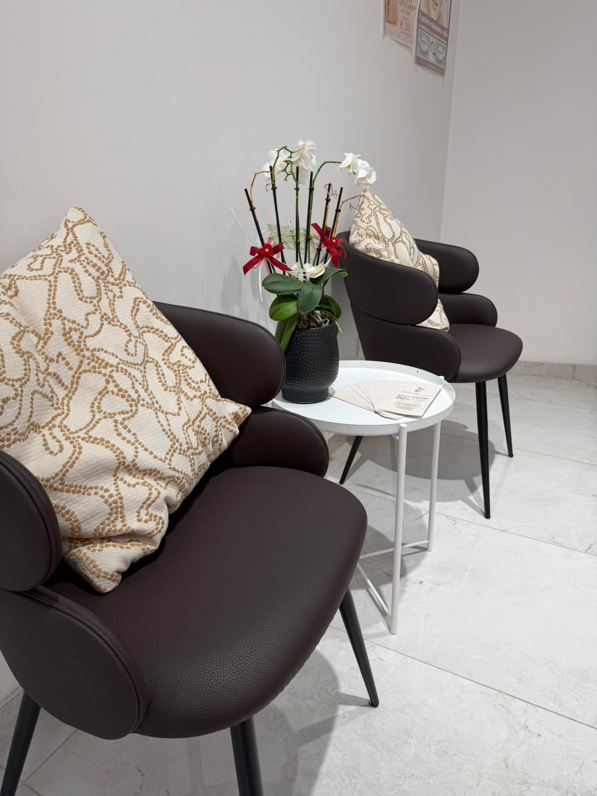 In una sala d'attesa, poltrone lounge marrone scuro con cuscini beige a fantasia, accanto a un tavolino bianco e una fioriera per orchidee.