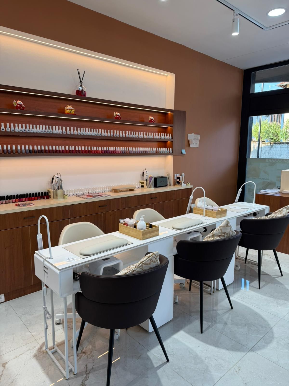 Moderno salone di manicure con tavoli bianchi, sedie nere e scaffali con prodotti sulla parete marrone.