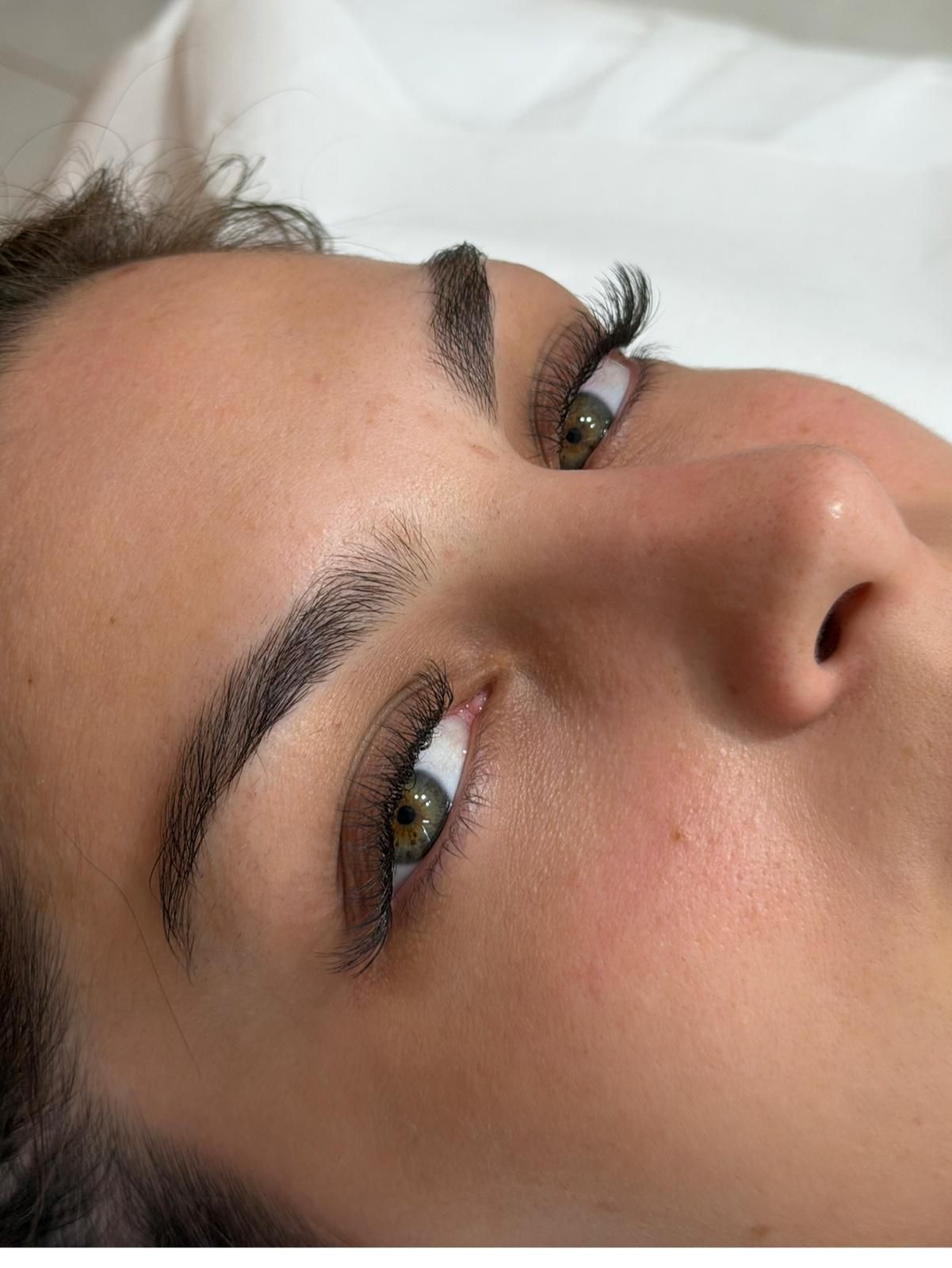 Primo piano del volto di una persona con occhi verdi, sopracciglia scure e pelle nuda, appoggiato su una superficie bianca.
