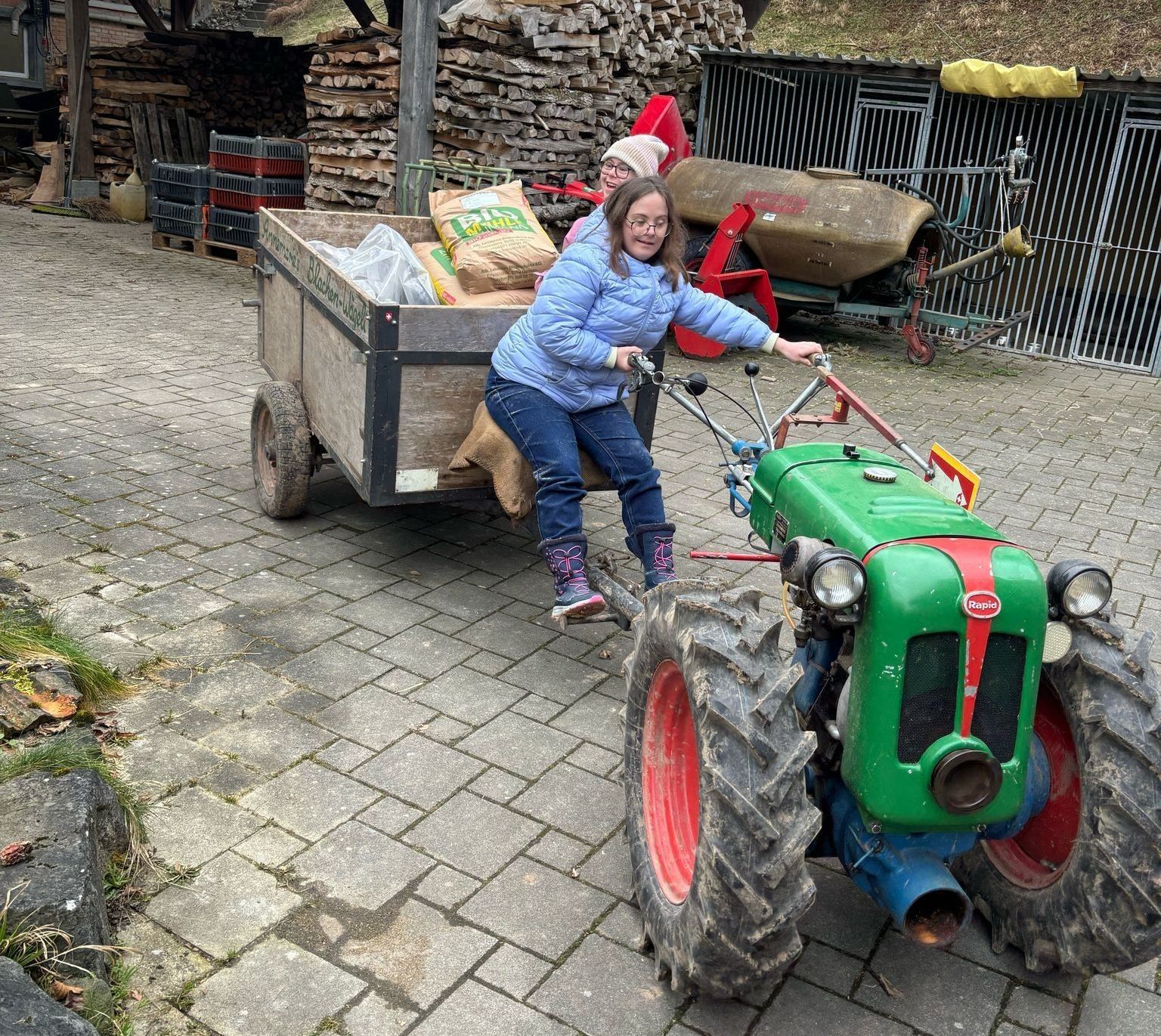 Eine Frau fährt einen kleinen grünen Traktor, der einen mit Säcken beladenen Anhänger zieht. Der Traktor steht auf einem gepflasterten Platz.