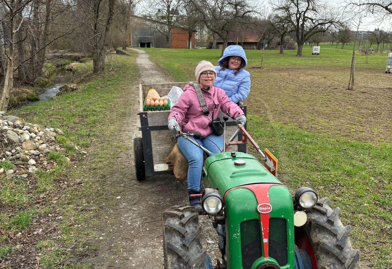 Zwei Personen auf einem grünen Traktor, der einen Wagen mit Eiern zieht, fahren auf einem Weg.