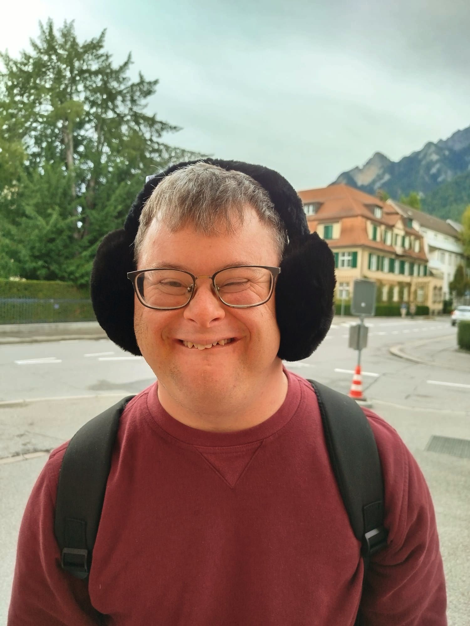 Mann mit Down-Syndrom und Ohrenschützern lächelt vor Gebäuden und Bäumen.