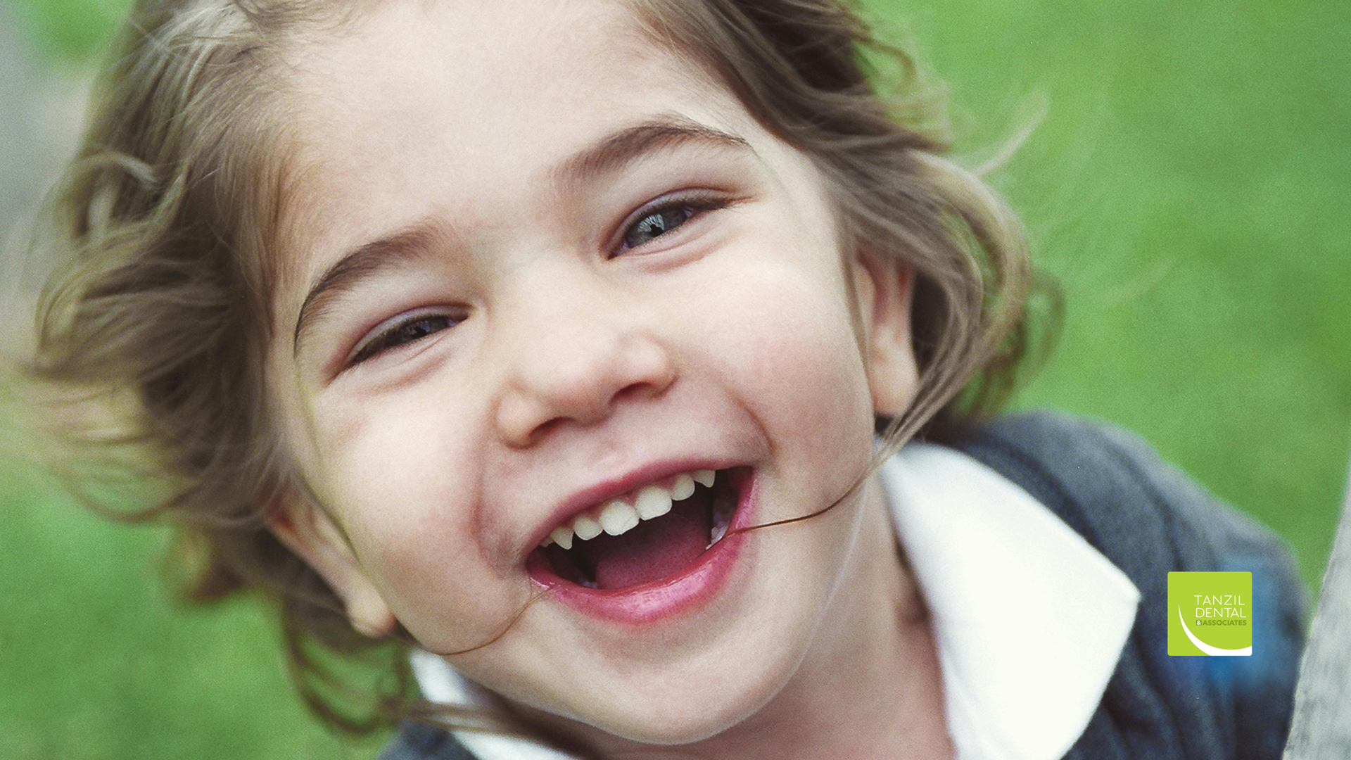 Smiling child with open mouth outdoors, with blurred green background.