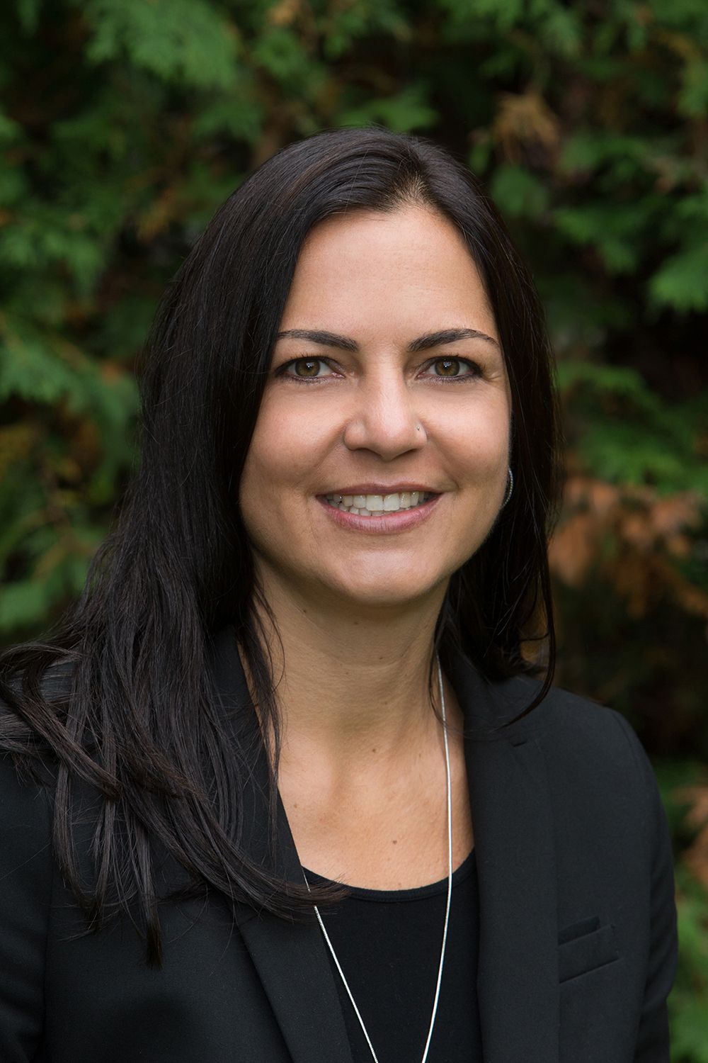 Woman in a black blazer smiles, stands outdoors in front of green foliage.