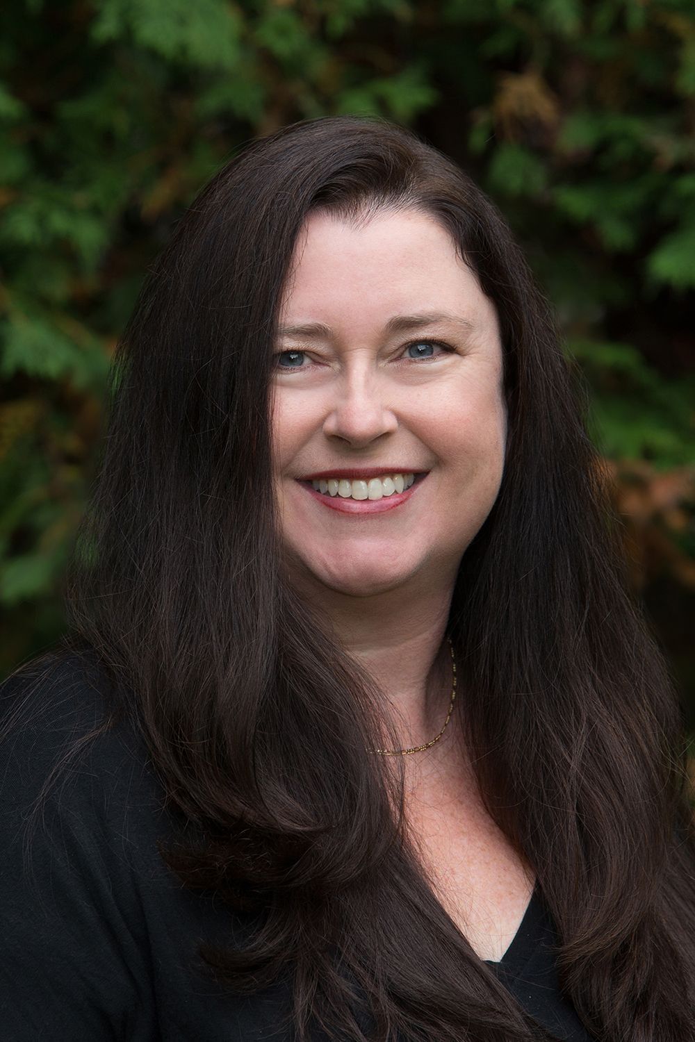 Woman with long, dark hair smiles outdoors, wearing black. Green background.
