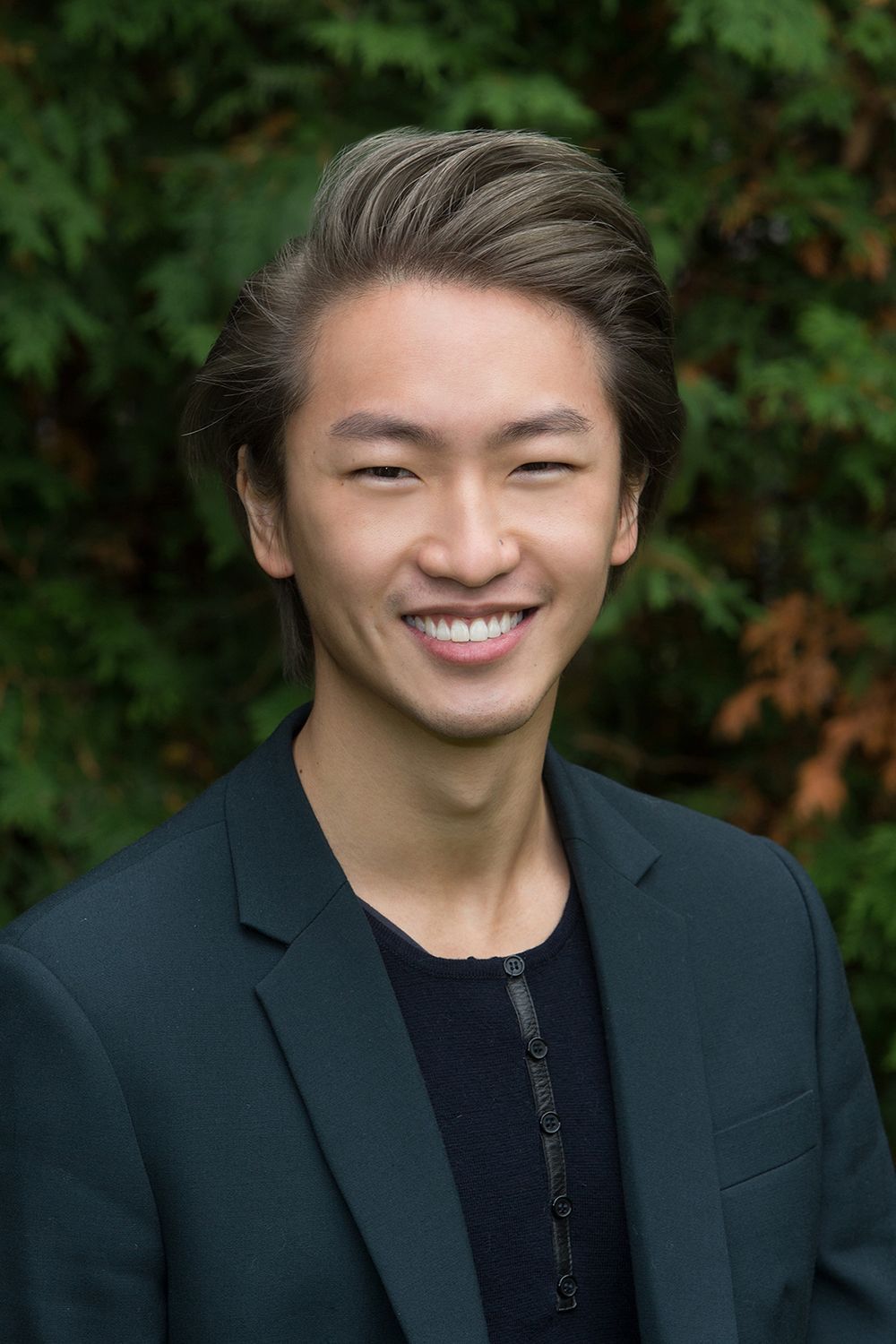 Smiling man with styled hair, wearing a dark blazer over a textured black shirt, set outdoors with green foliage.