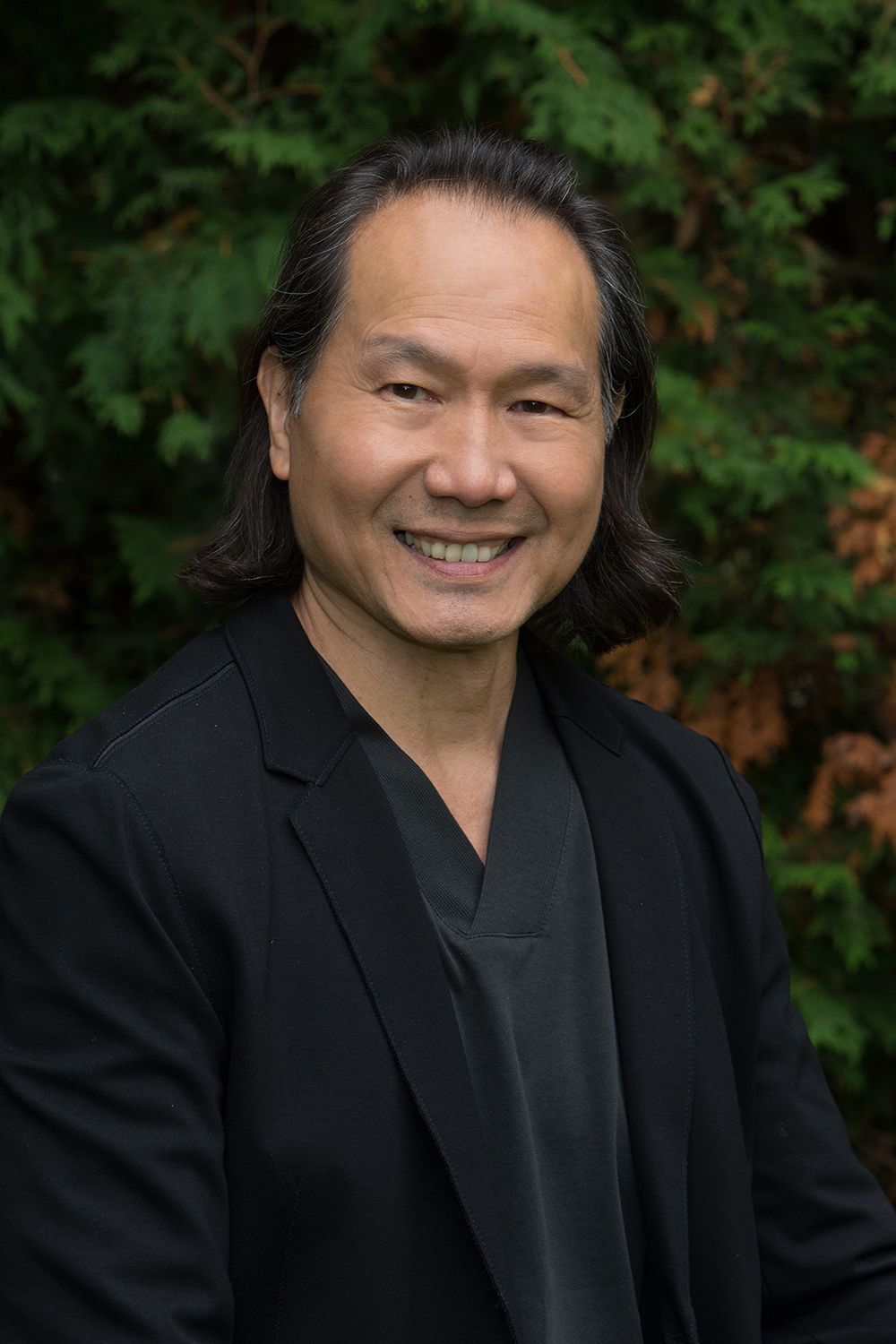 Man with long dark hair, smiling, wearing a black blazer and gray top, set against a green and brown background.