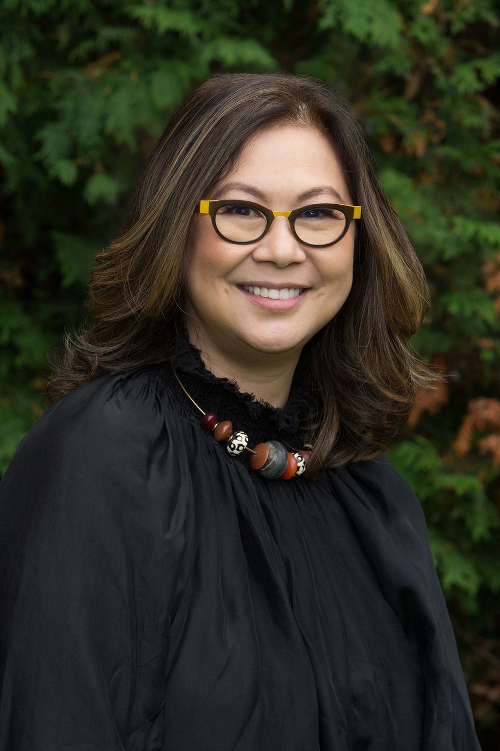 Woman with glasses and dark hair smiles, wearing a black top and beaded necklace, set in front of green foliage.