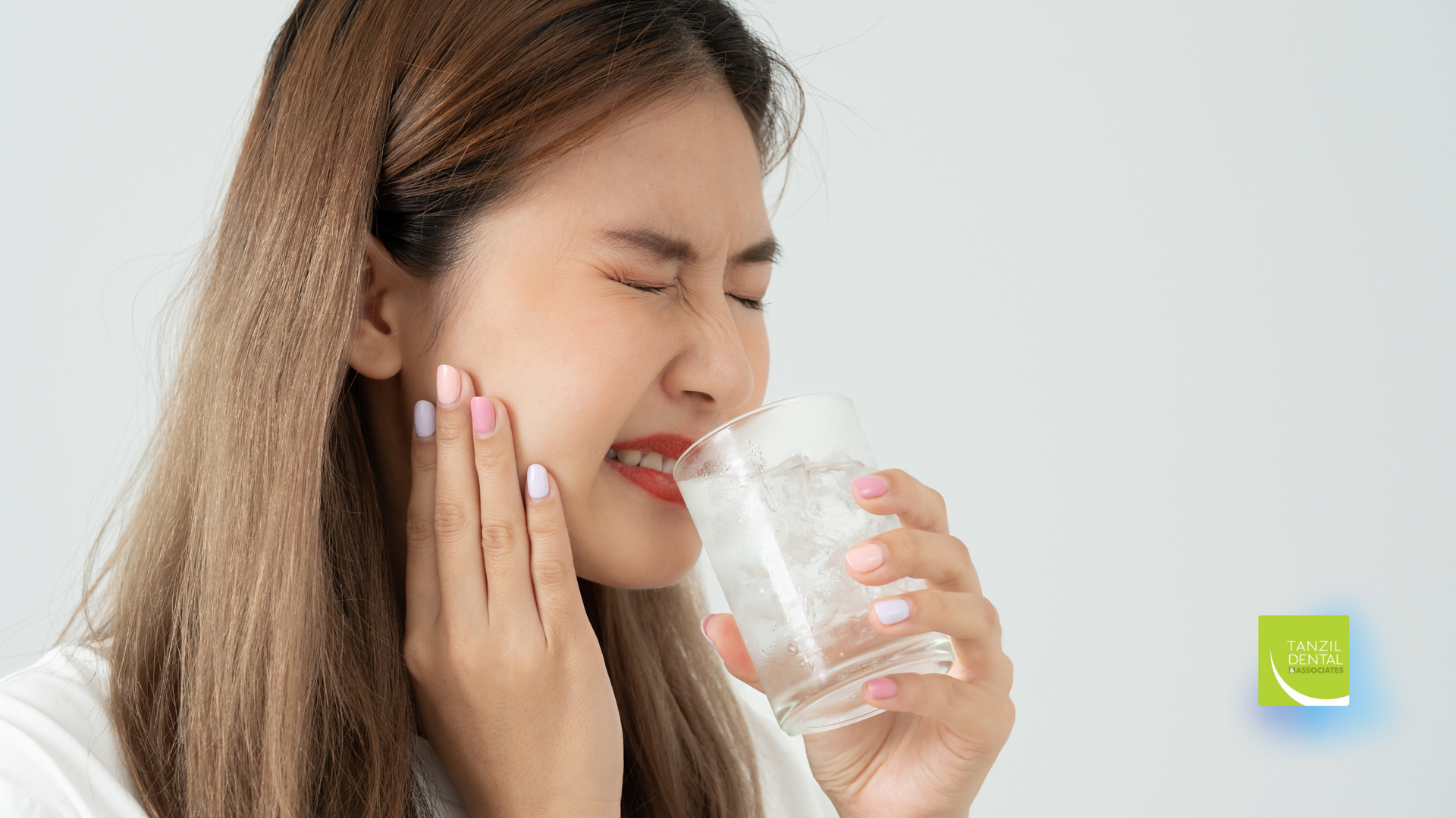 A woman is drinking a glass of water while holding her mouth in pain.