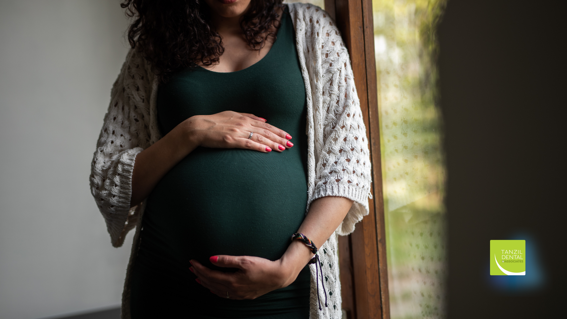 A pregnant woman is standing in front of a window holding her belly.