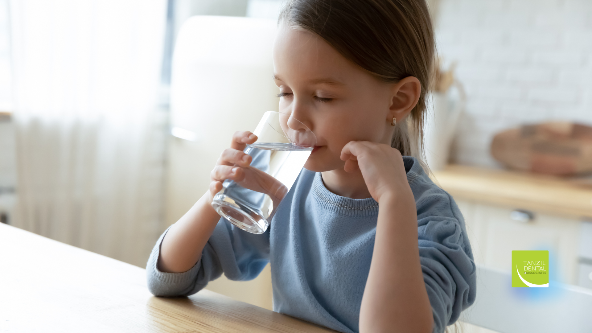 A young girl drinking water from a clear glass, sitting at a table indoors.