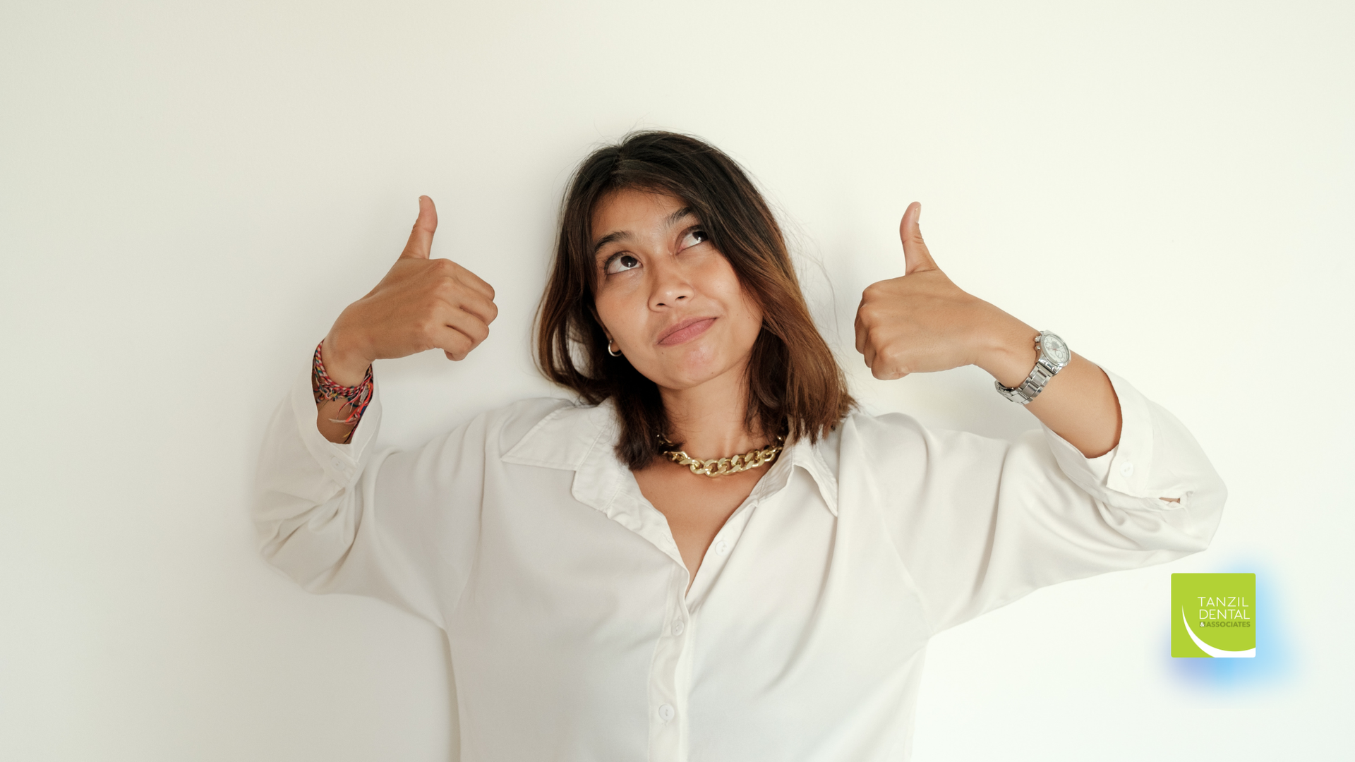 Woman in white shirt giving thumbs up, looking upward, against a white wall.