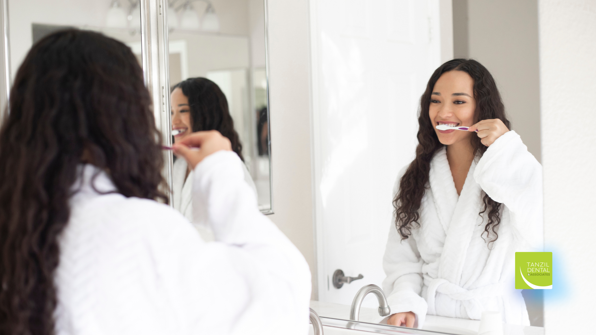 Woman in white robe brushing teeth in front of a bathroom mirror. Smiling with a toothbrush.