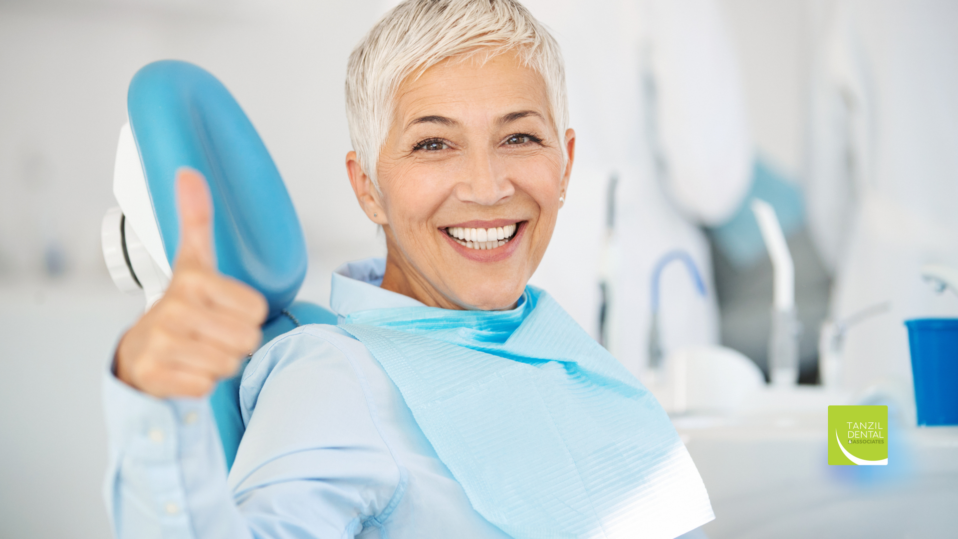 An older woman is sitting in a dental chair and giving a thumbs up.