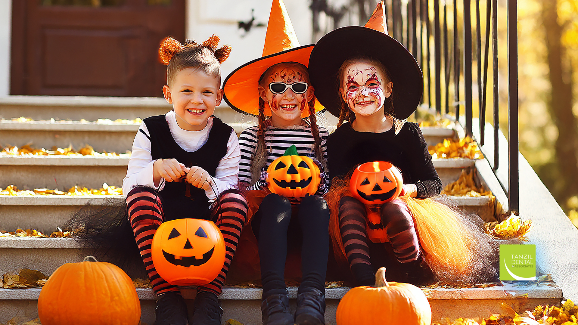 Three children in Halloween costumes on steps, holding jack-o'-lanterns; fall foliage in background.