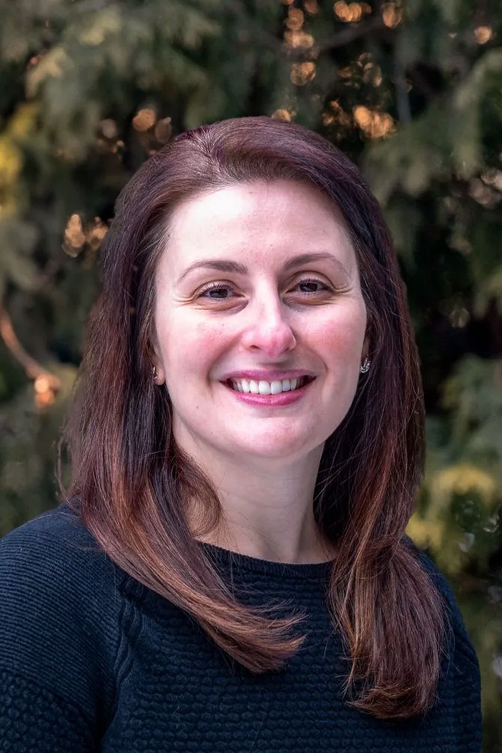 Woman with brown hair smiles, wearing a black sweater, set against a blurred green background.