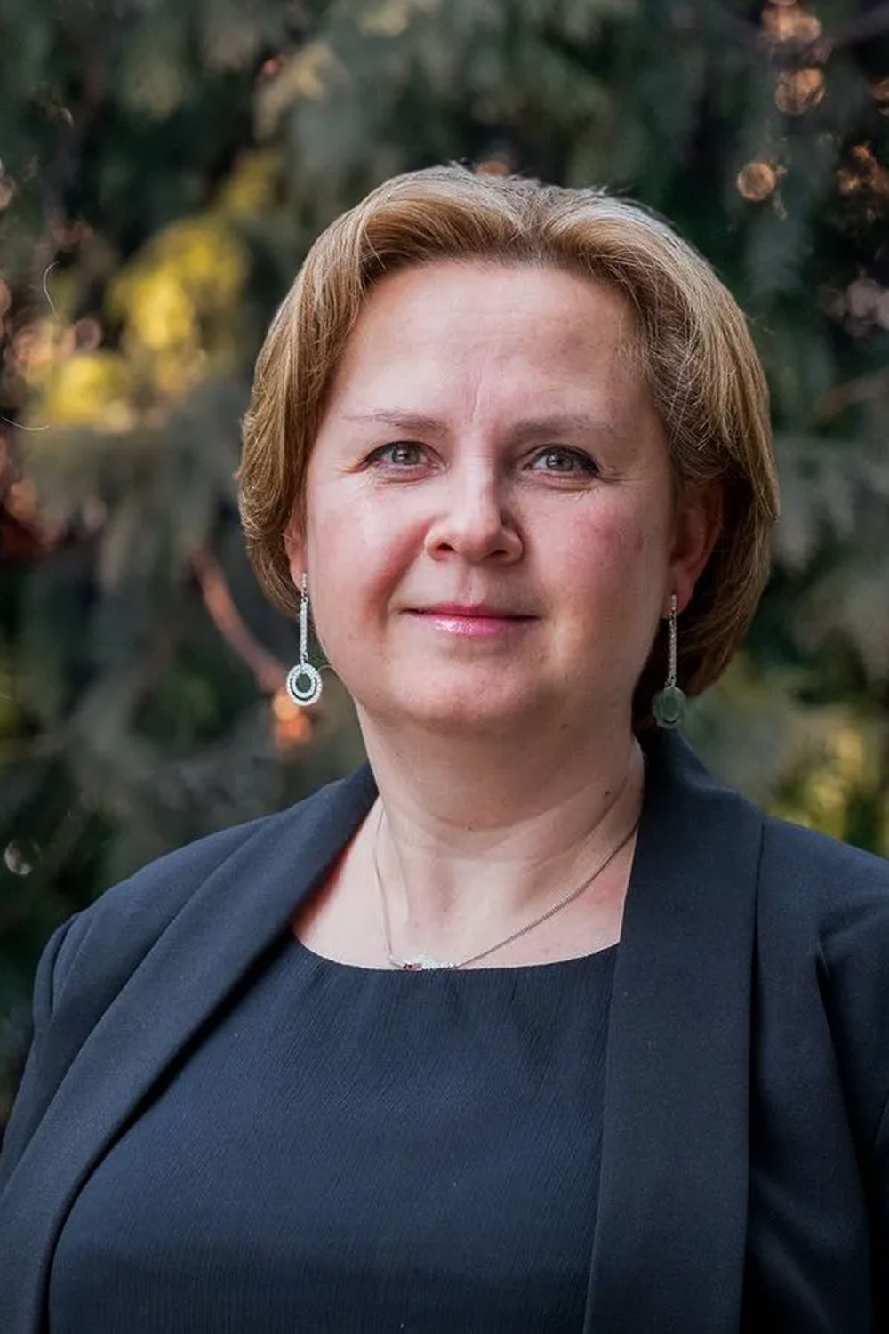 Woman with short blonde hair, wearing a dark blazer and earrings, smiling.