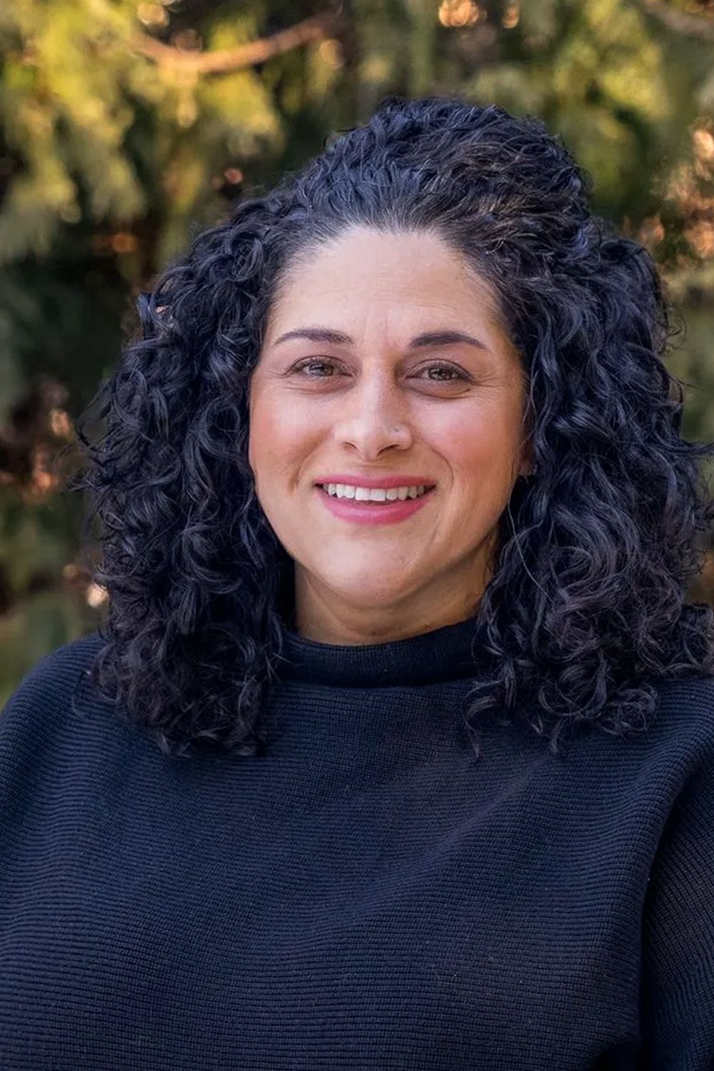 Woman with dark curly hair smiles at the camera, wearing a dark sweater; outdoors.