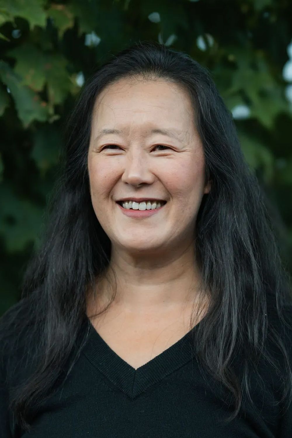 Woman with long, dark hair smiles. Wearing a black top, set outdoors with green foliage in the background.