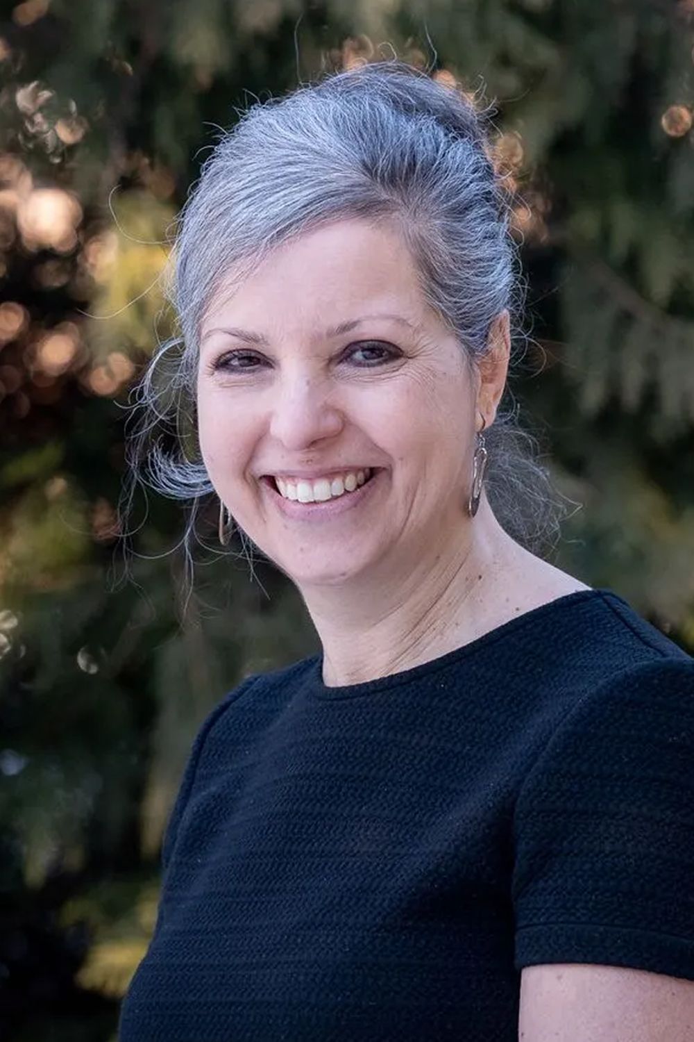 Woman with gray hair smiles, wearing a black top, outdoors with blurred green background.