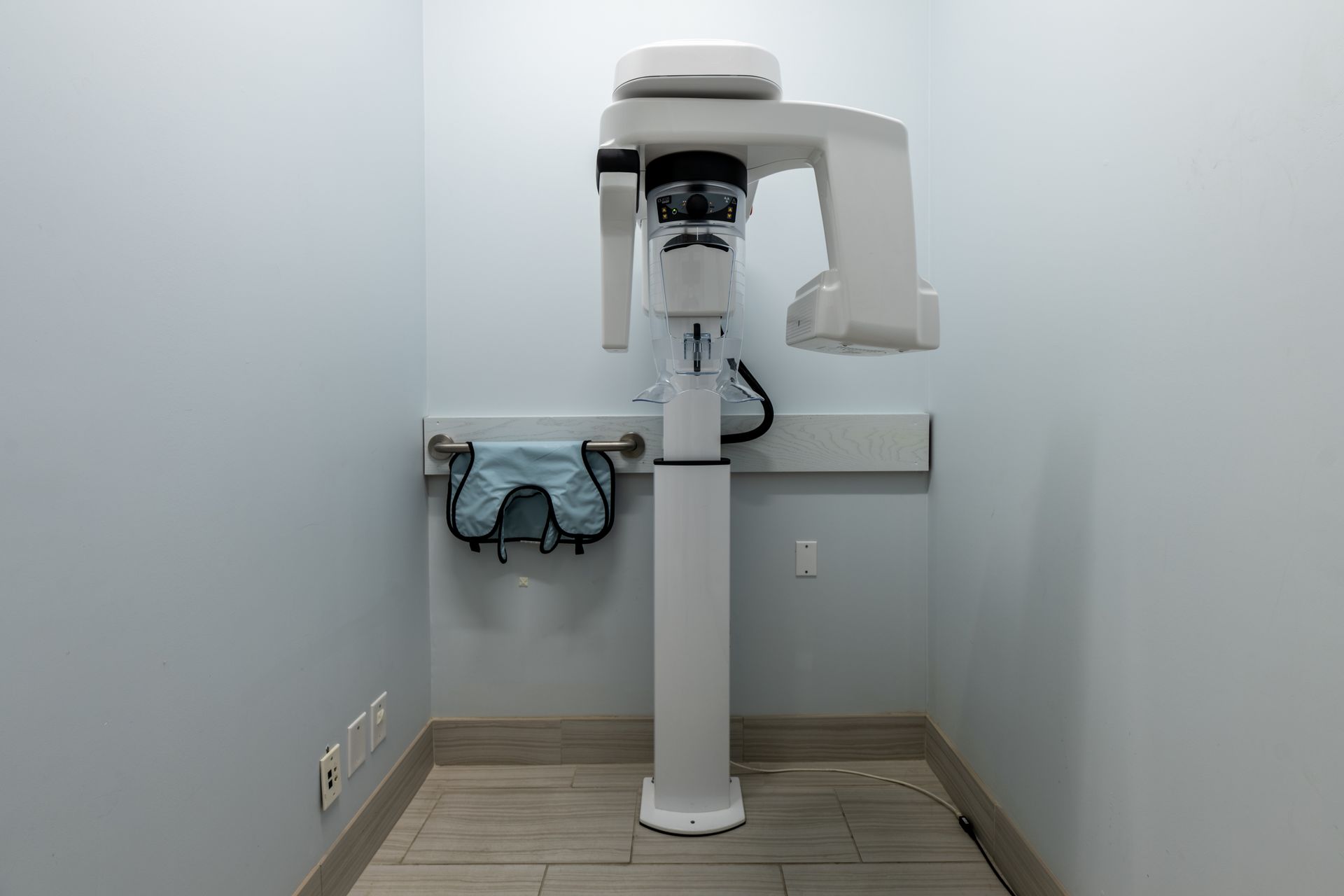 Panoramic dental X-ray machine in a small room with a lead apron hanging nearby.