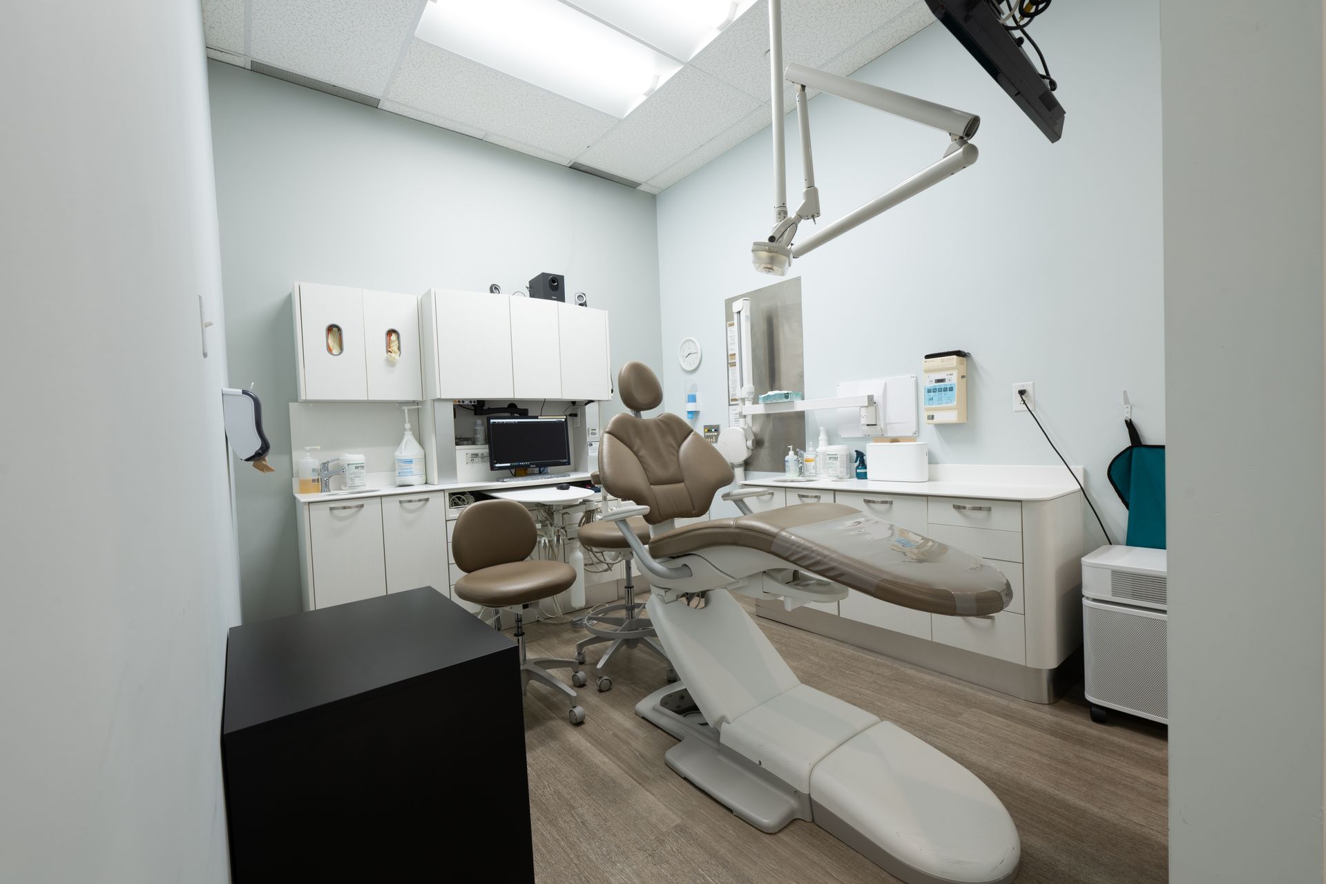 Dental exam room with chair, cabinets, and equipment; light blue walls, wood floor.