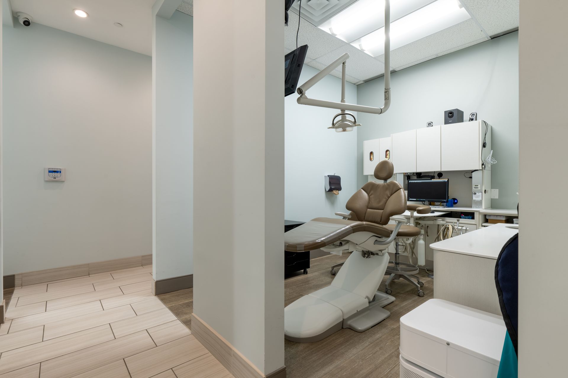 Dental office interior: hallway leads to a dental exam room with equipment and chair, light blue walls, overhead light.