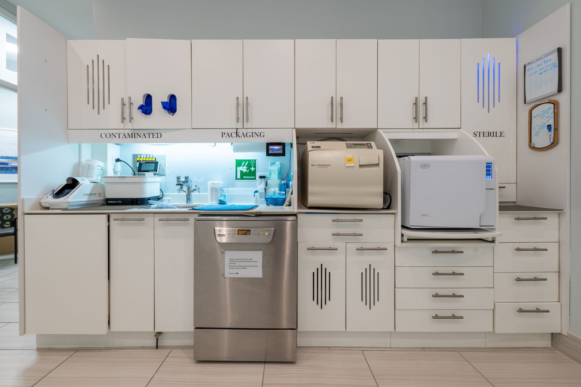 Dental sterilization equipment and storage cabinets. White cabinets, stainless steel appliances, and safety signage.