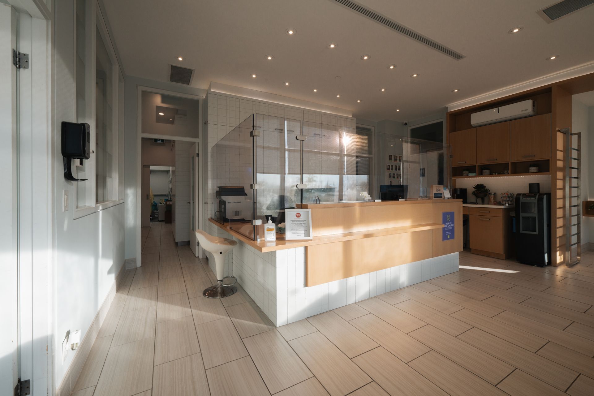 Bright reception area with white and wood tones; a counter with a protective barrier and coffee equipment.