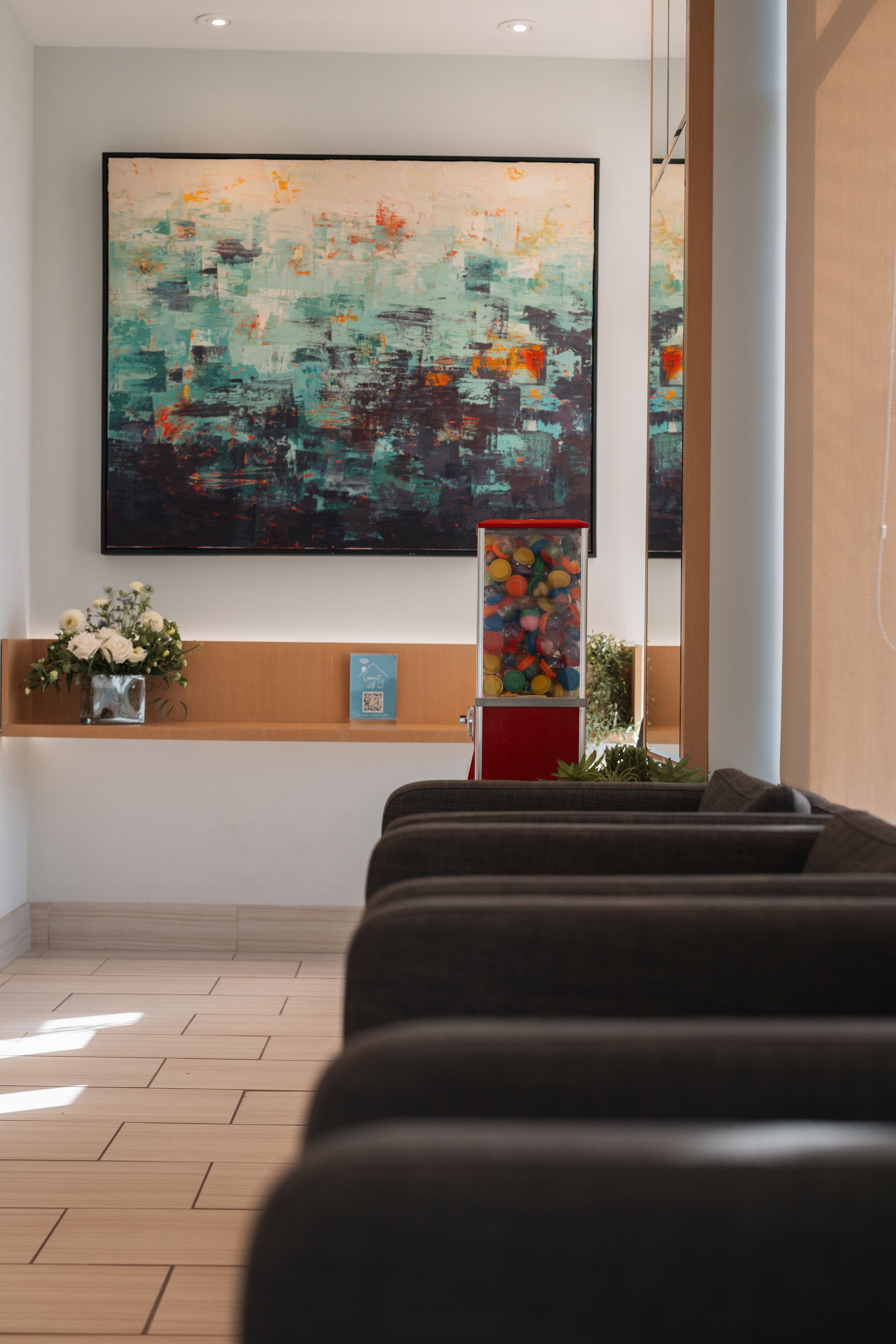 Lobby with abstract art, gumball machine, and a row of dark seating. Flowers and books are on a wooden shelf.