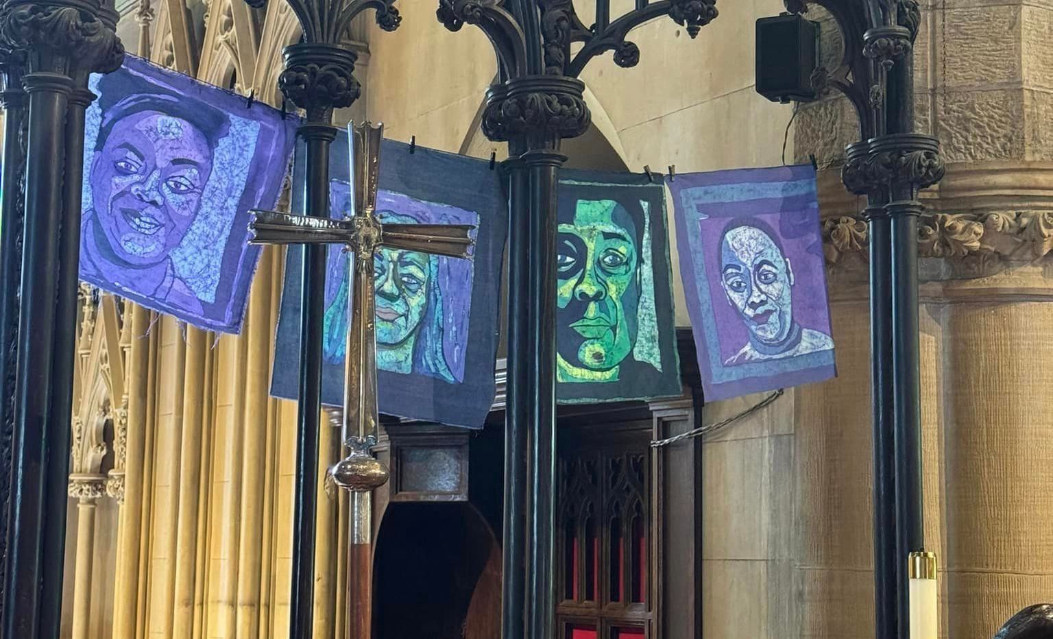 Four colorful artistic portraits hanging in a row inside an ornate, stone-columned church, positioned behind a metal cross.
