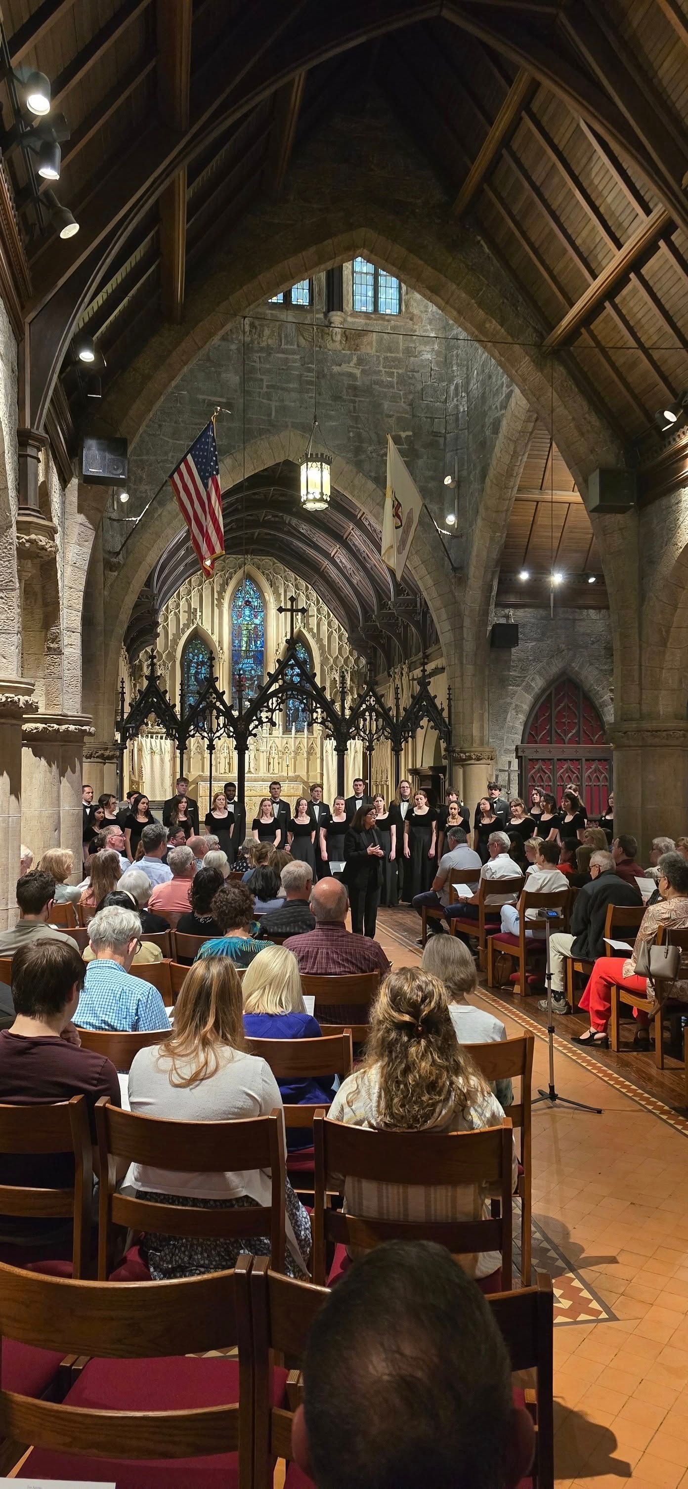 A choir performs on an altar in a stone cathedral, with a seated audience viewing them from wooden pews.
