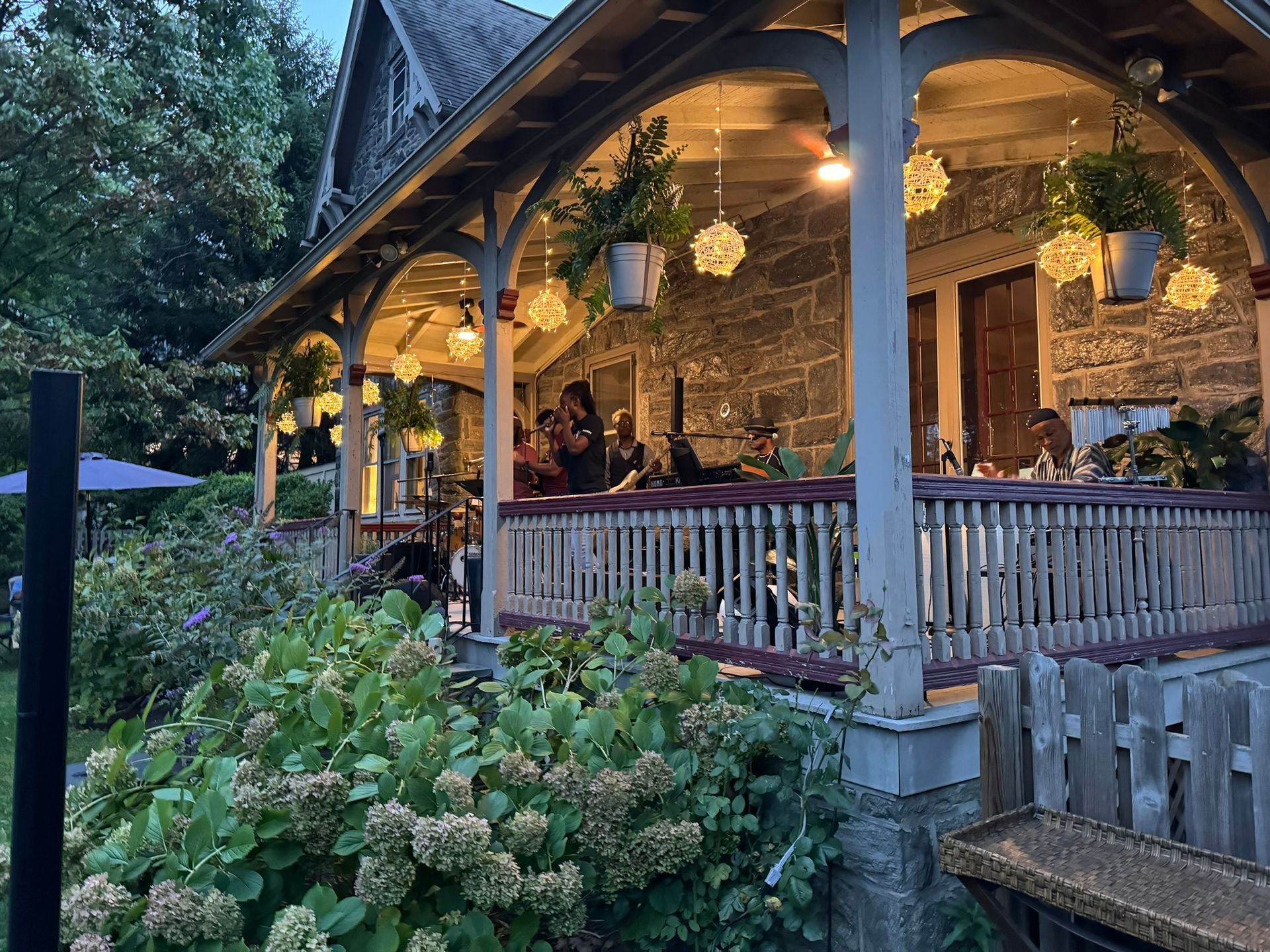A stone cottage porch with hanging plants and warm lights, featuring people dining at tables on a wooden deck.