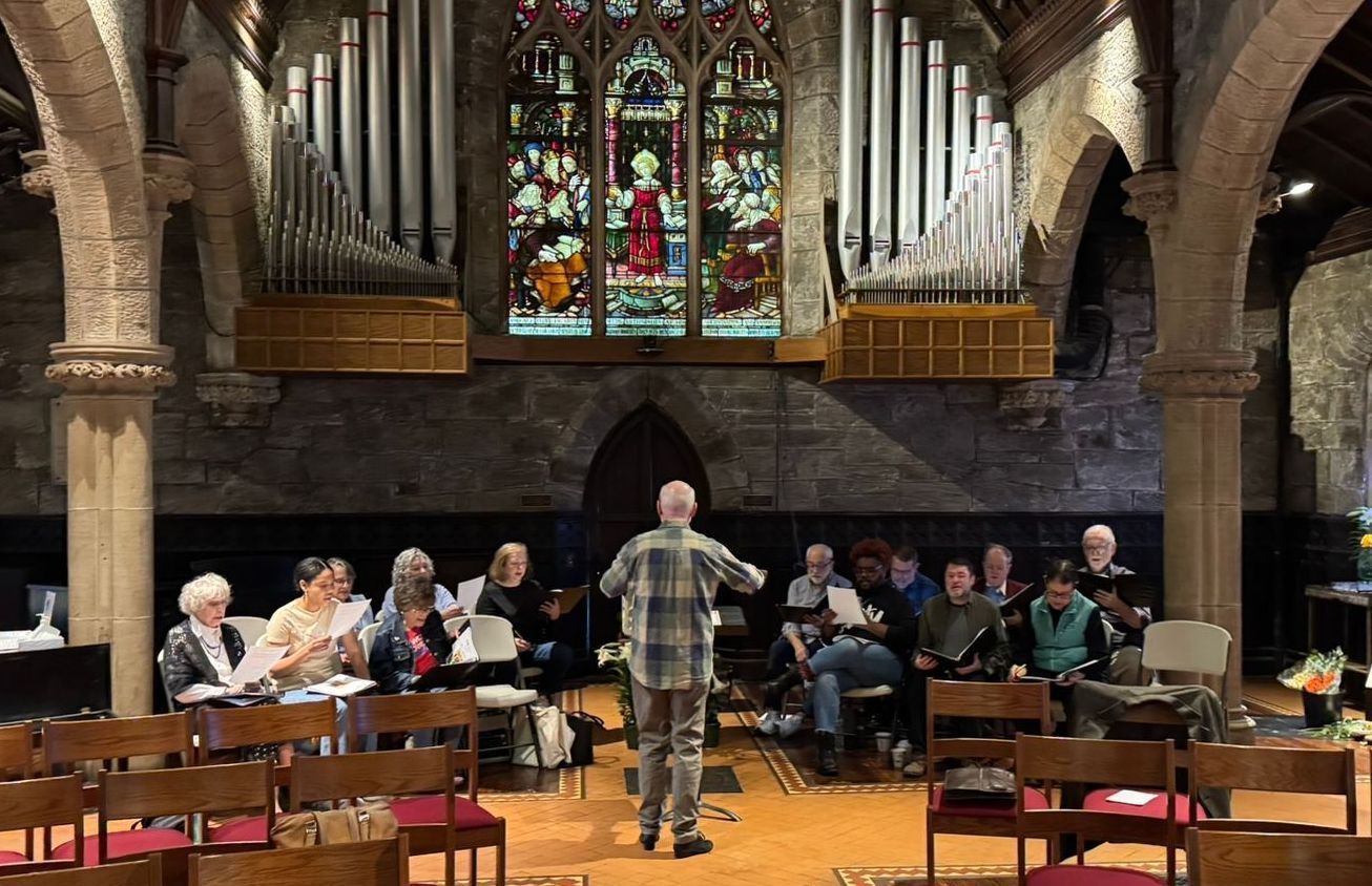A choir rehearses in a stone church, led by a conductor standing with their back to the camera.