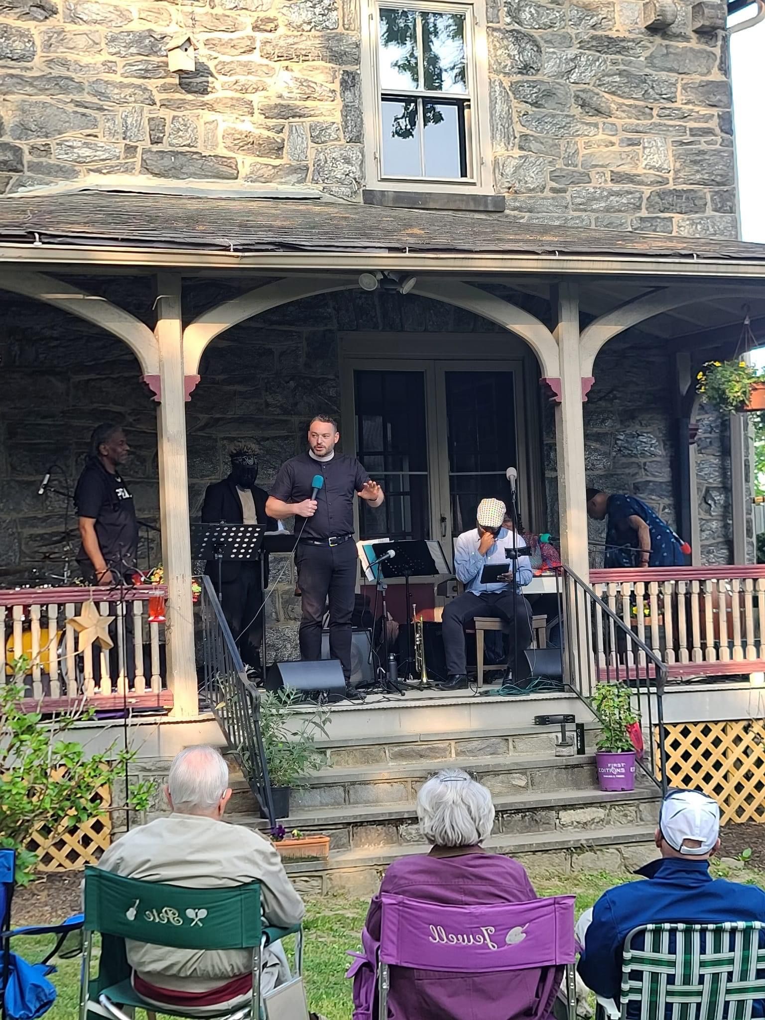A small band plays music on the front porch of a stone building for a seated audience on a sunny day.