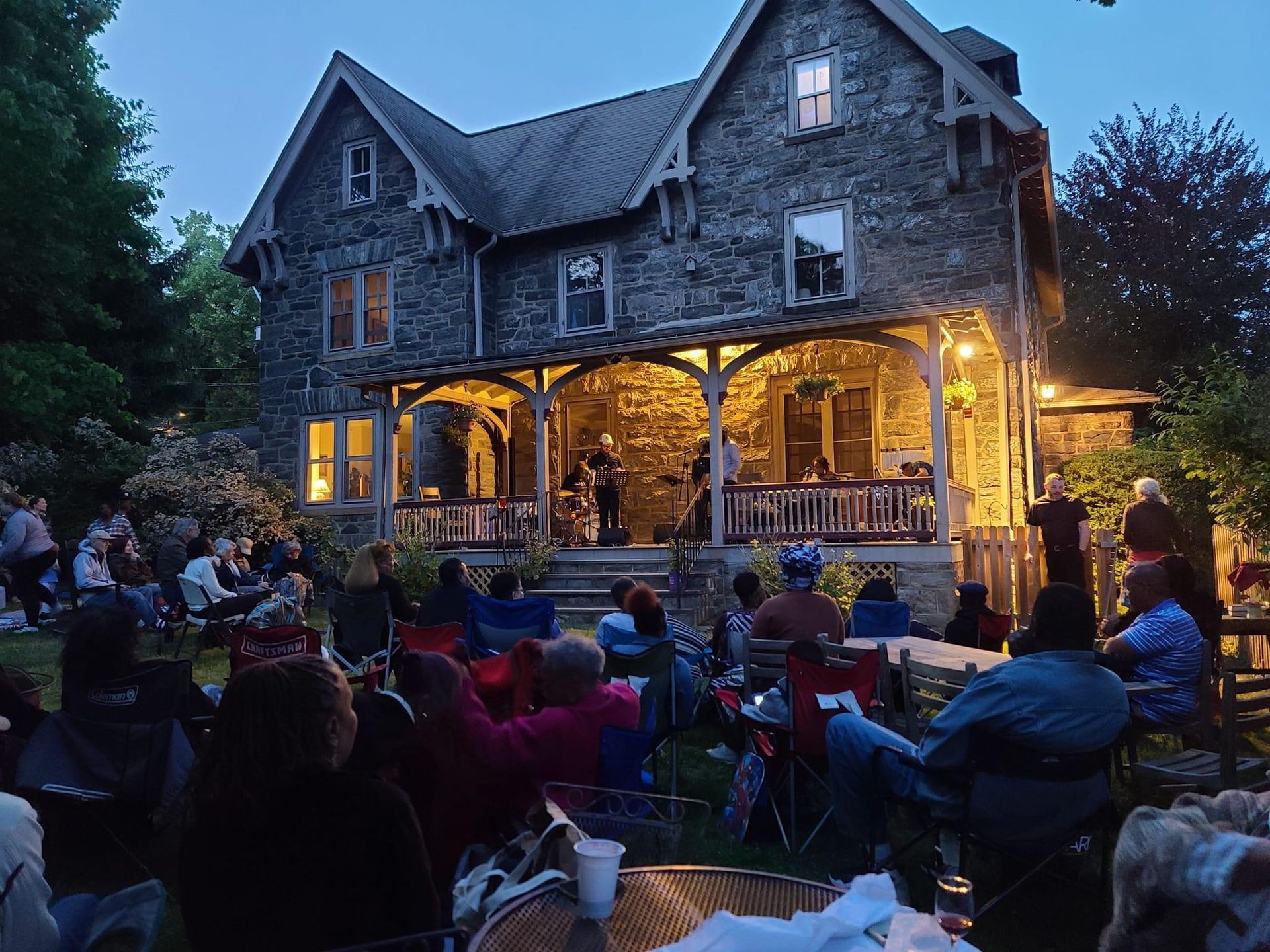 People watch a live band perform on the lighted porch of a historic stone house at dusk.