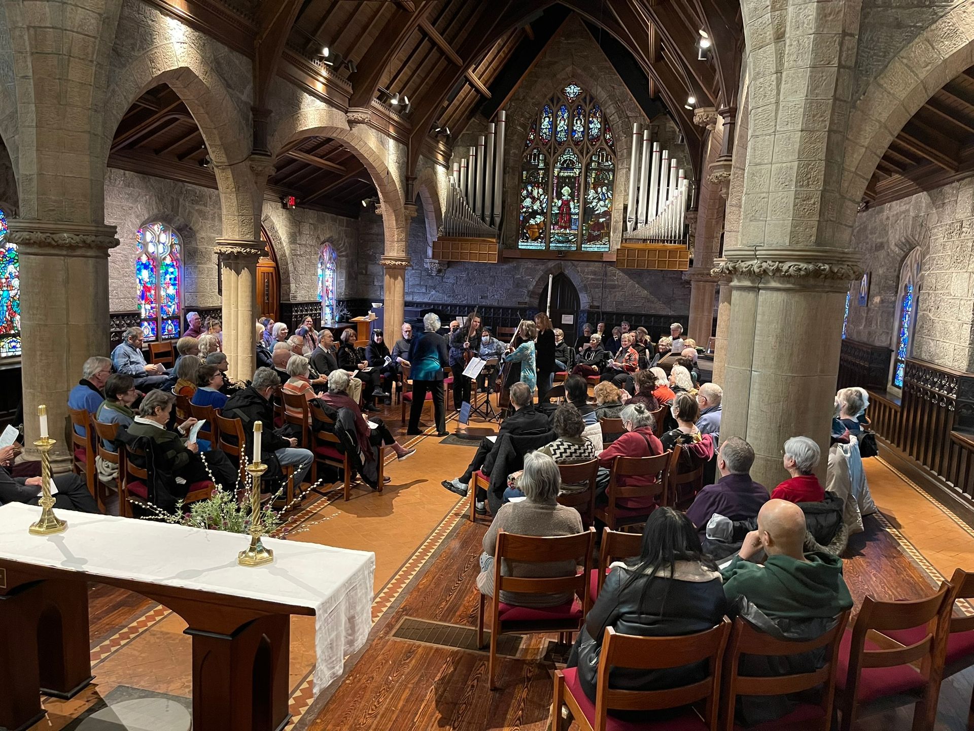 A church interior with a gathered audience seated on wooden chairs facing a performance area near a stained-glass window.