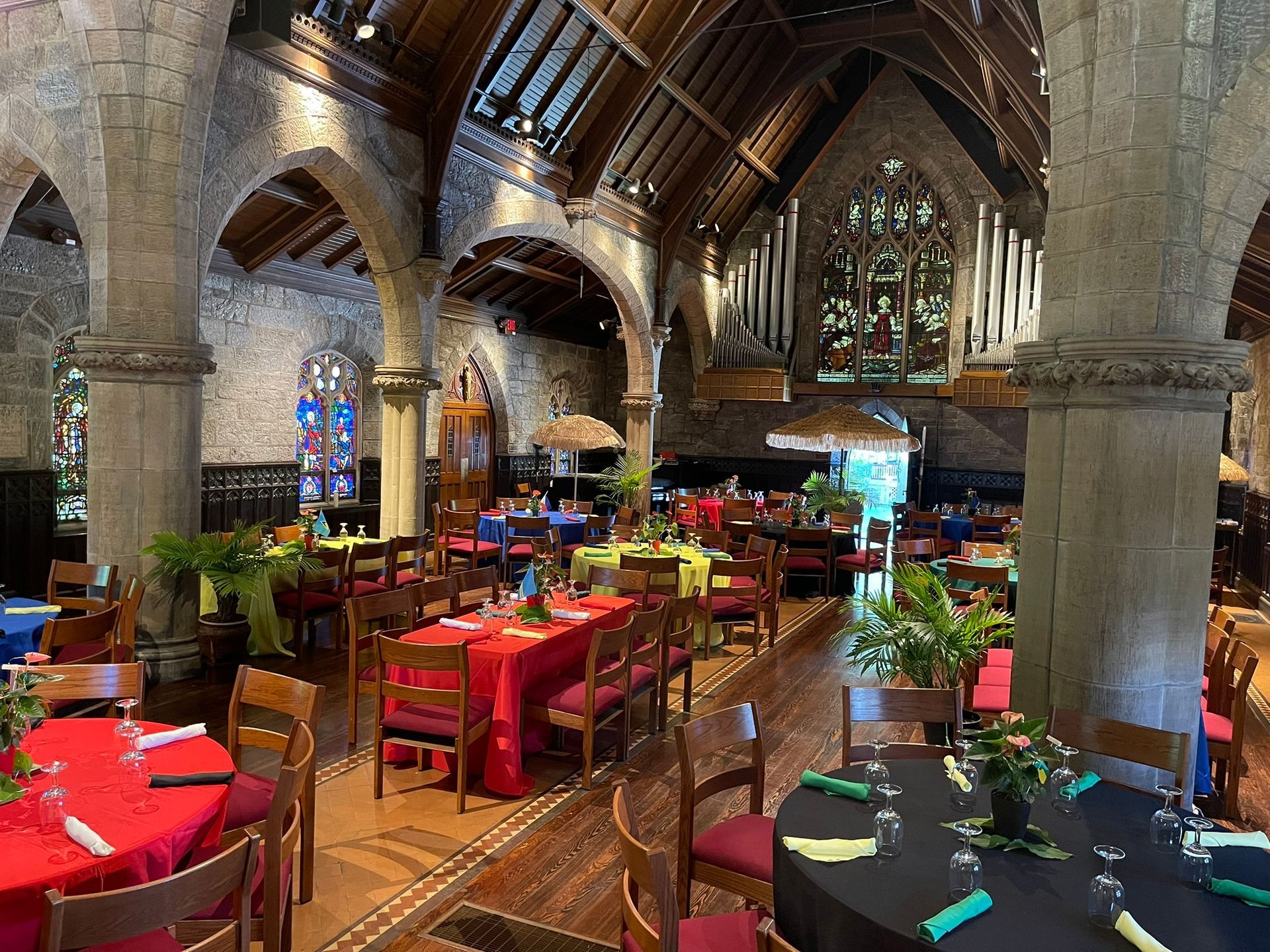 A rustic hall with stone pillars, vaulted ceilings, and tables set with vibrant red and black tablecloths for an event.