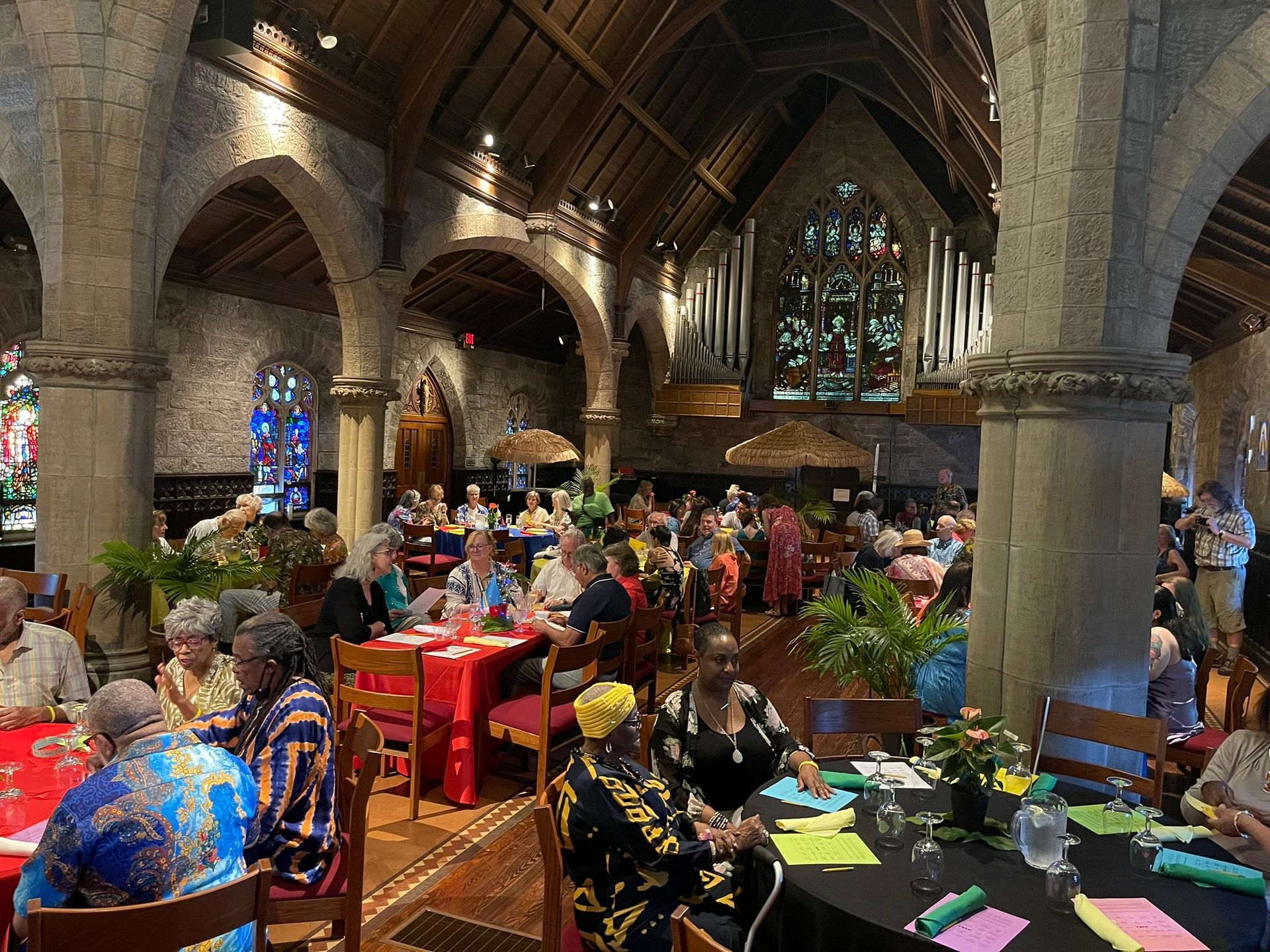 People dine at round tables in a decorated, high-ceilinged hall featuring stone columns and stained glass windows.