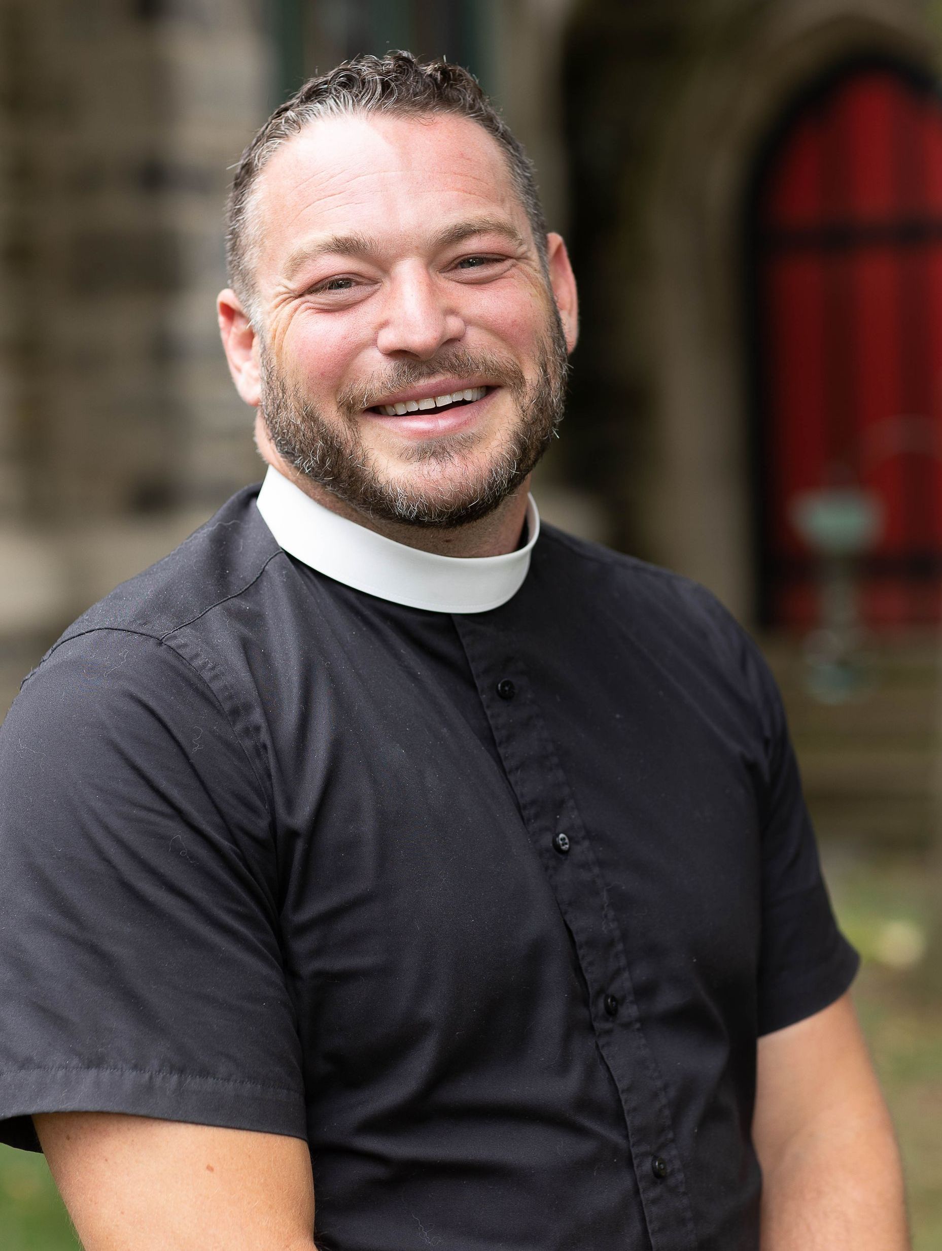 A smiling person wearing a black clerical shirt with a white clerical collar, standing in front of a stone building.