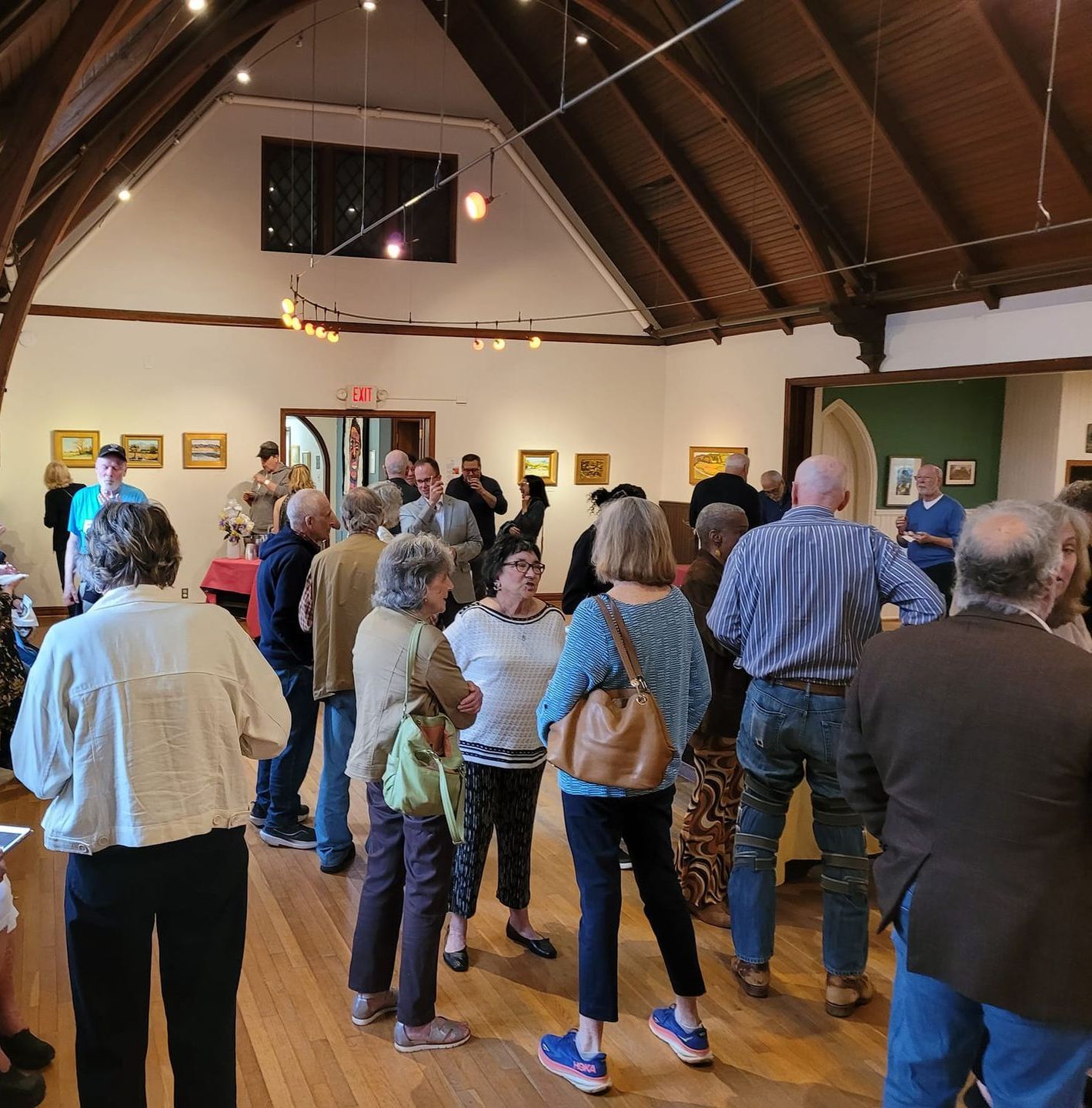 A diverse group of people gathers in a vaulted gallery space with framed art on the walls to socialize.