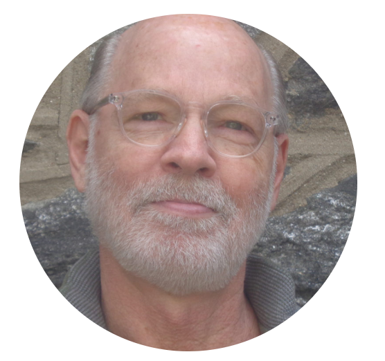 Circular headshot of an individual with a gray beard and glasses, posing against a stone wall background.