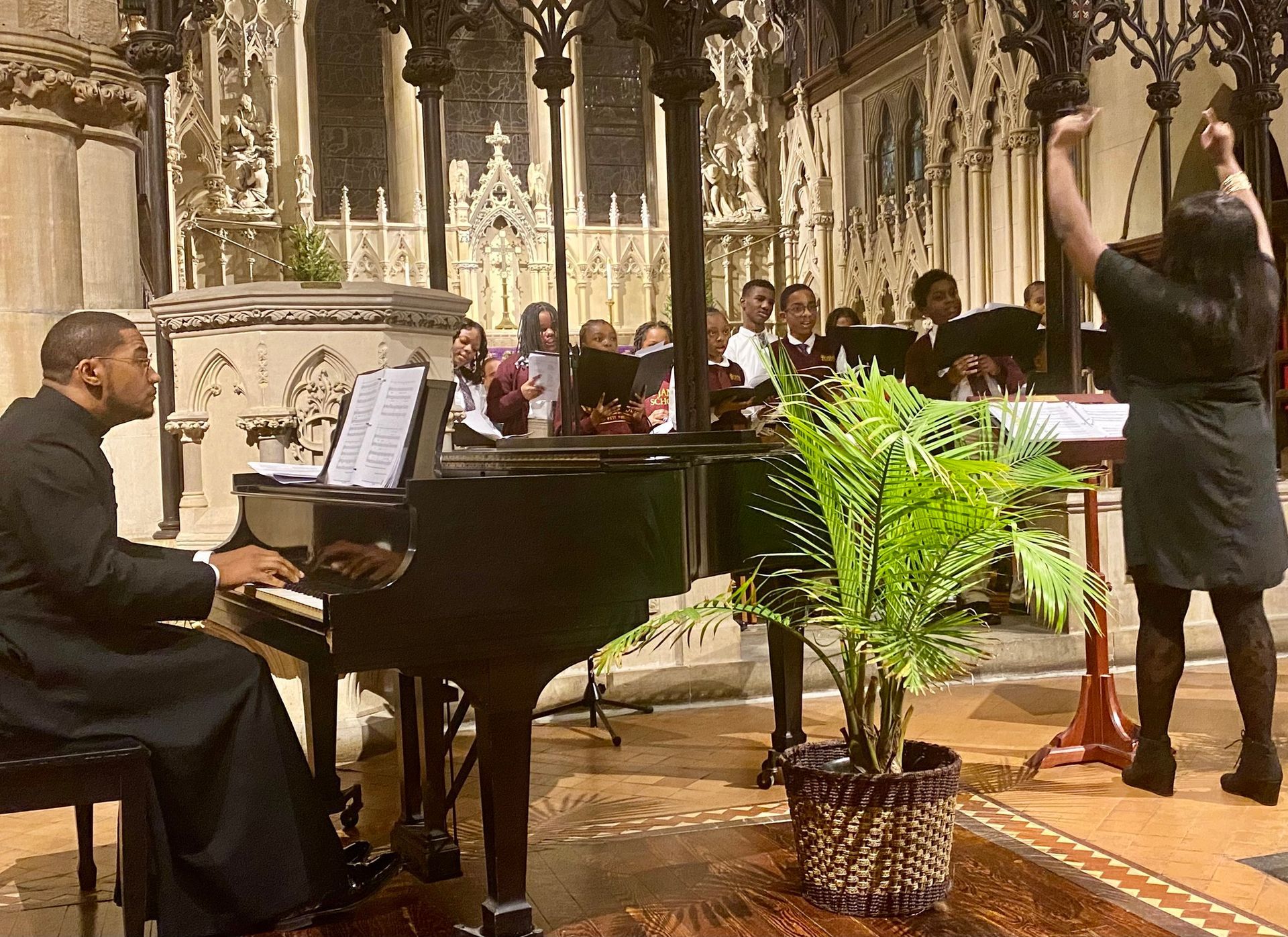 A pianist plays a grand piano inside a church, accompanied by a choir in the background and a conductor leading them.