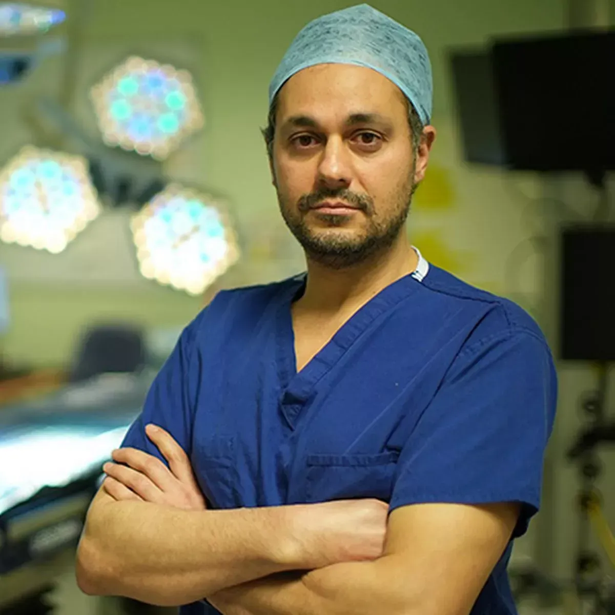 Surgeon in blue scrubs and cap standing with arms crossed in an operating room