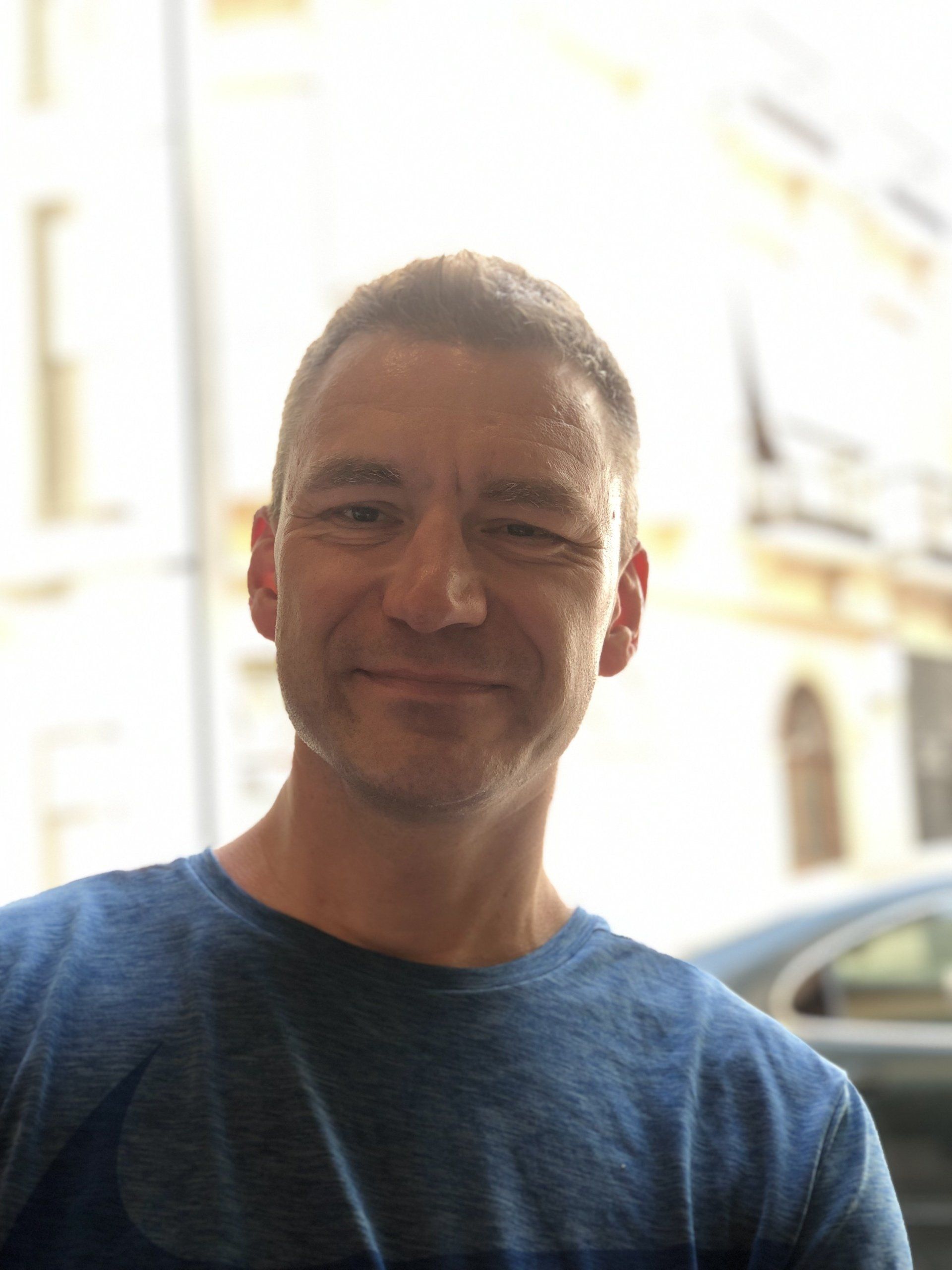 Smiling man in a blue shirt standing outdoors in bright sunlight
